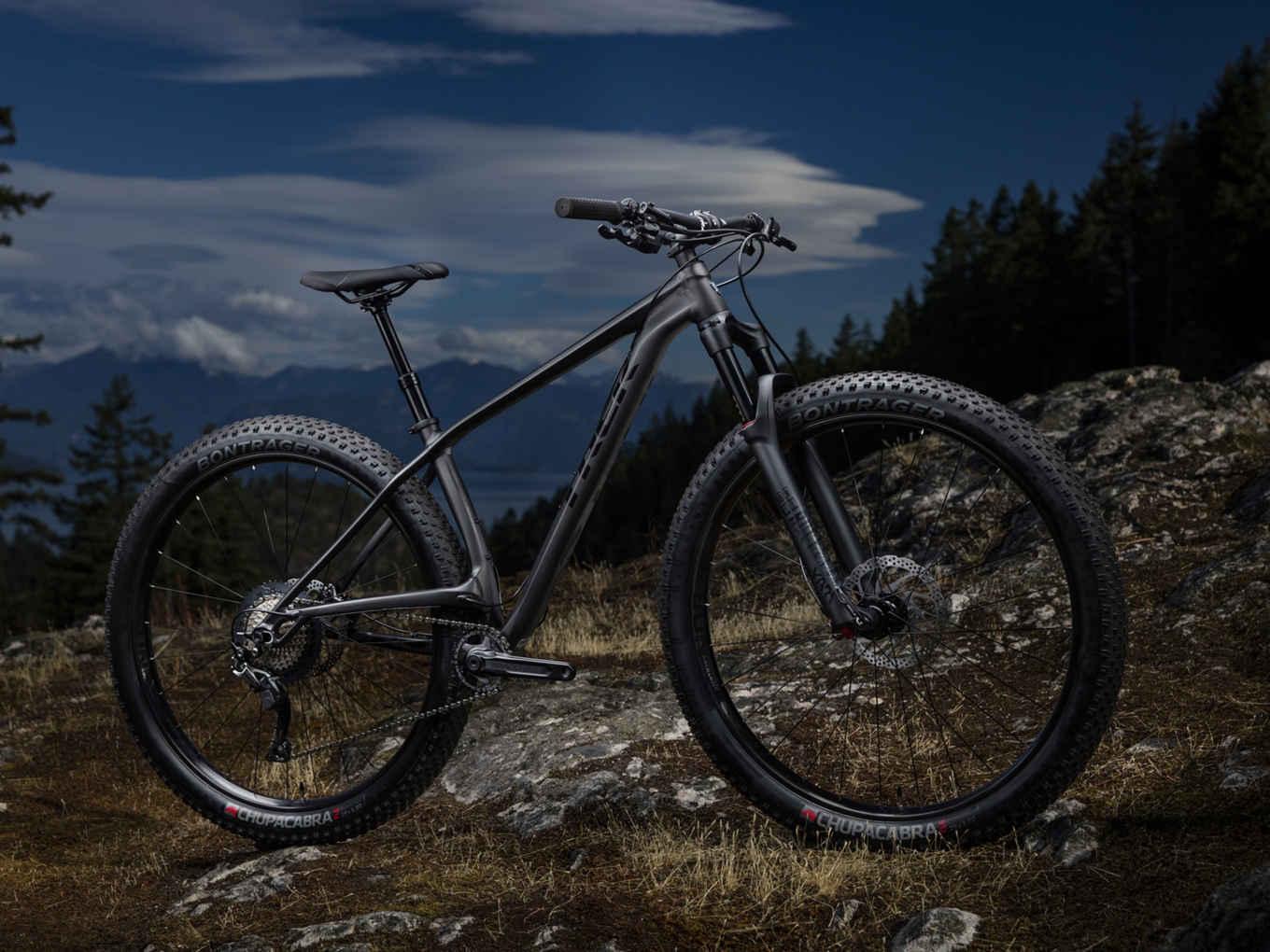 Trek Stache: A modern mountain bike positioned on rocky terrain, showcasing a sleek design with a matte gray frame. The bike features wide, knobby tires suitable for off-road riding and is set against a backdrop of lush trees and a dramatic sky with clouds.