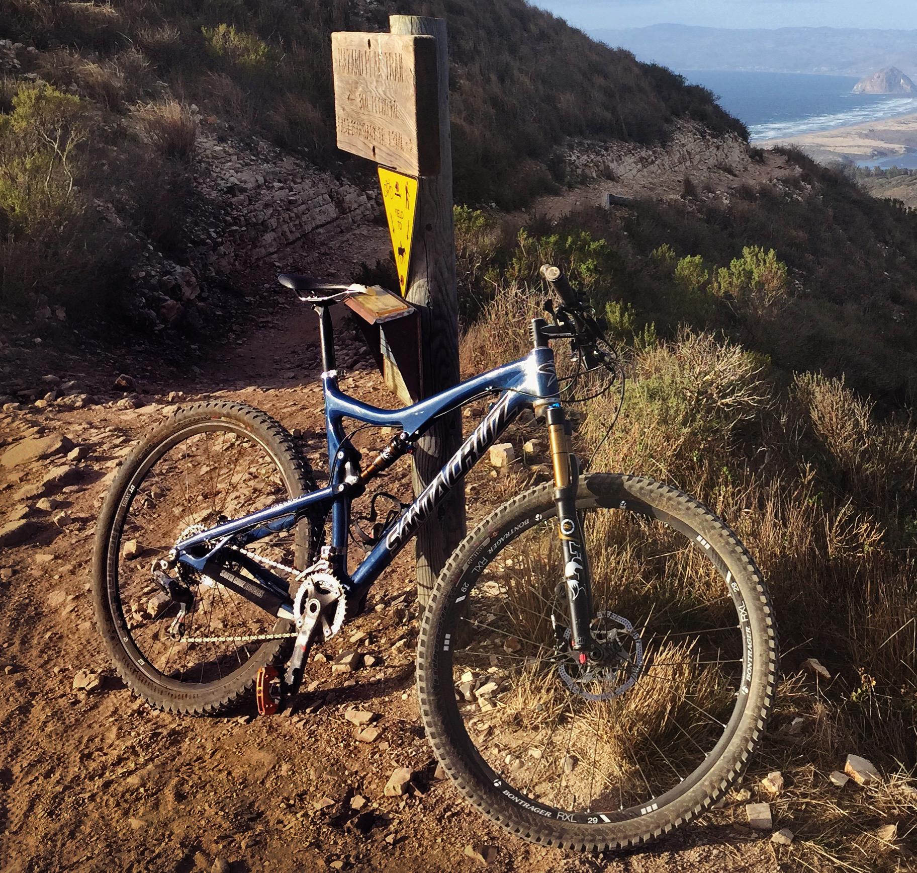 Santa Cruz Tallboy Carbon: A mountain bike resting near a trail sign on a rocky path, surrounded by vegetation and overlooking a scenic landscape with hills and coastline in the distance.