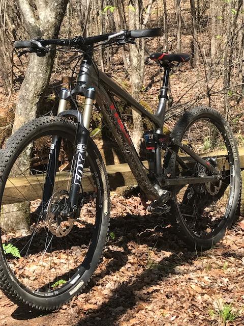 Trek Fuel EX 8: A mountain bike resting on a bed of fallen leaves, positioned near a wooden fence in a forested area. The bike features black and silver components, including thick tires designed for off-road terrain and dual front suspension. Sunlight filters through the trees, highlighting the scene.