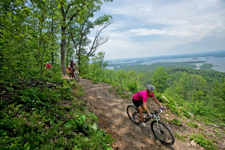 A woman rides a mountain bike along a dirt trail in a lush green forest, with two other cyclists visible in the background. The scene overlooks a vast landscape of trees and bodies of water, under a bright sky.
