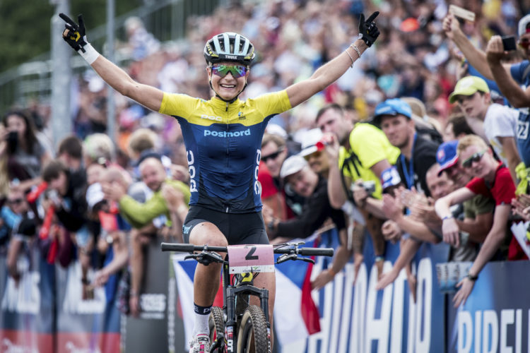 A mountain biker celebrating victory with arms raised in excitement, wearing a yellow and blue racing outfit. The backdrop features a cheering crowd of spectators and colorful banners, capturing the energy of the moment.
