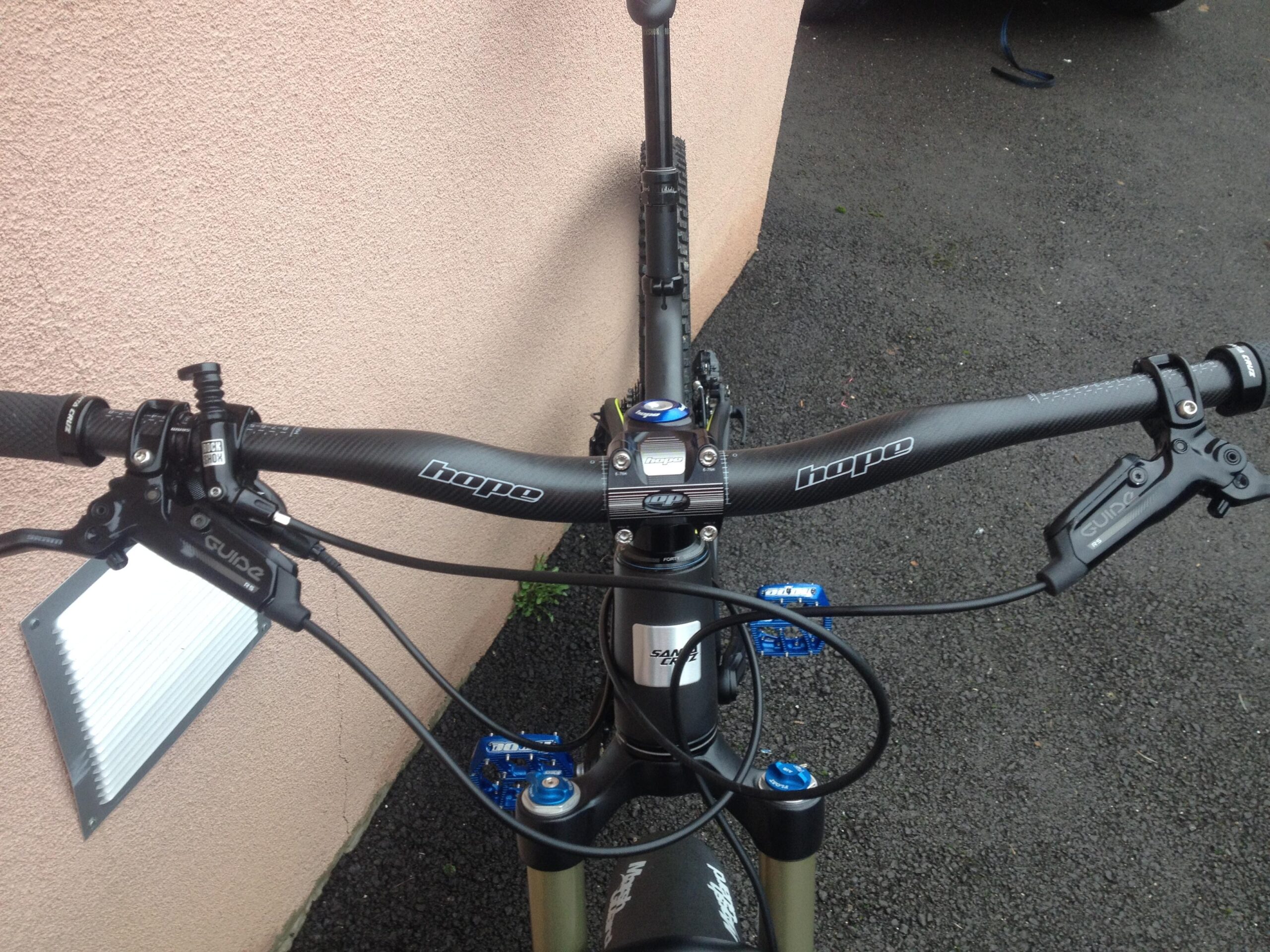 Santa Cruz Tallboy Carbon: Close-up view of a bicycle handlebar featuring a carbon fiber bar with "Hope" branding, hydraulic brake levers, and a brake cable. A silver bike light mount is visible on the left side. The background shows a textured pink wall and some black gravel surface.
