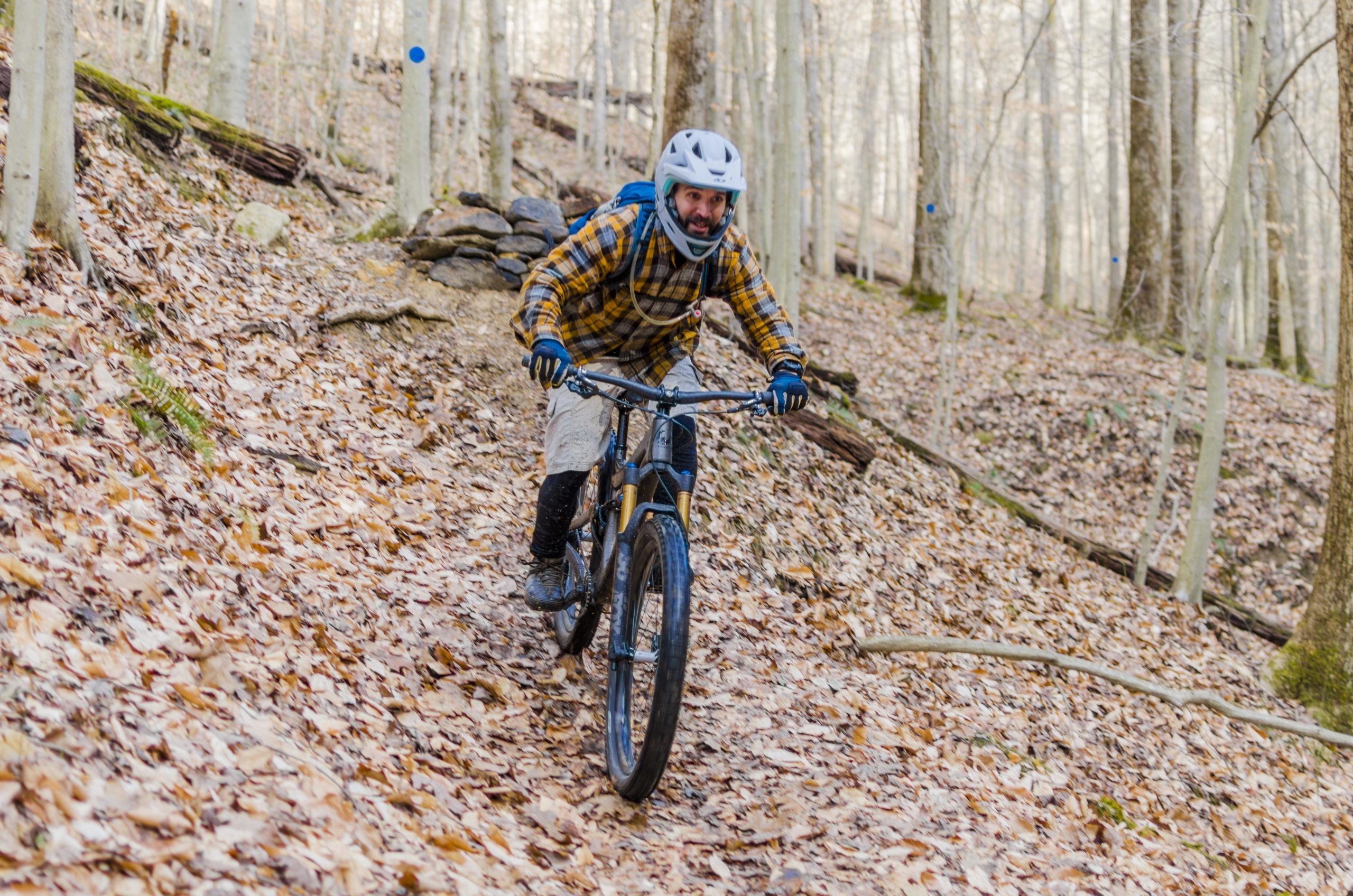 A mountain biker riding down a leaf-covered trail in a forest, wearing a helmet, gloves, and a plaid shirt. The background features bare trees and rocks along the path. Black Bear Trail mountain bike trail.