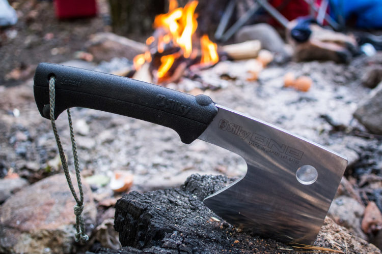 A silvery cleaver-style knife resting on a piece of wood, with a black rubber handle and a looped cord. In the background, a campfire blazes, surrounded by natural terrain and camping gear, creating an outdoor setting.