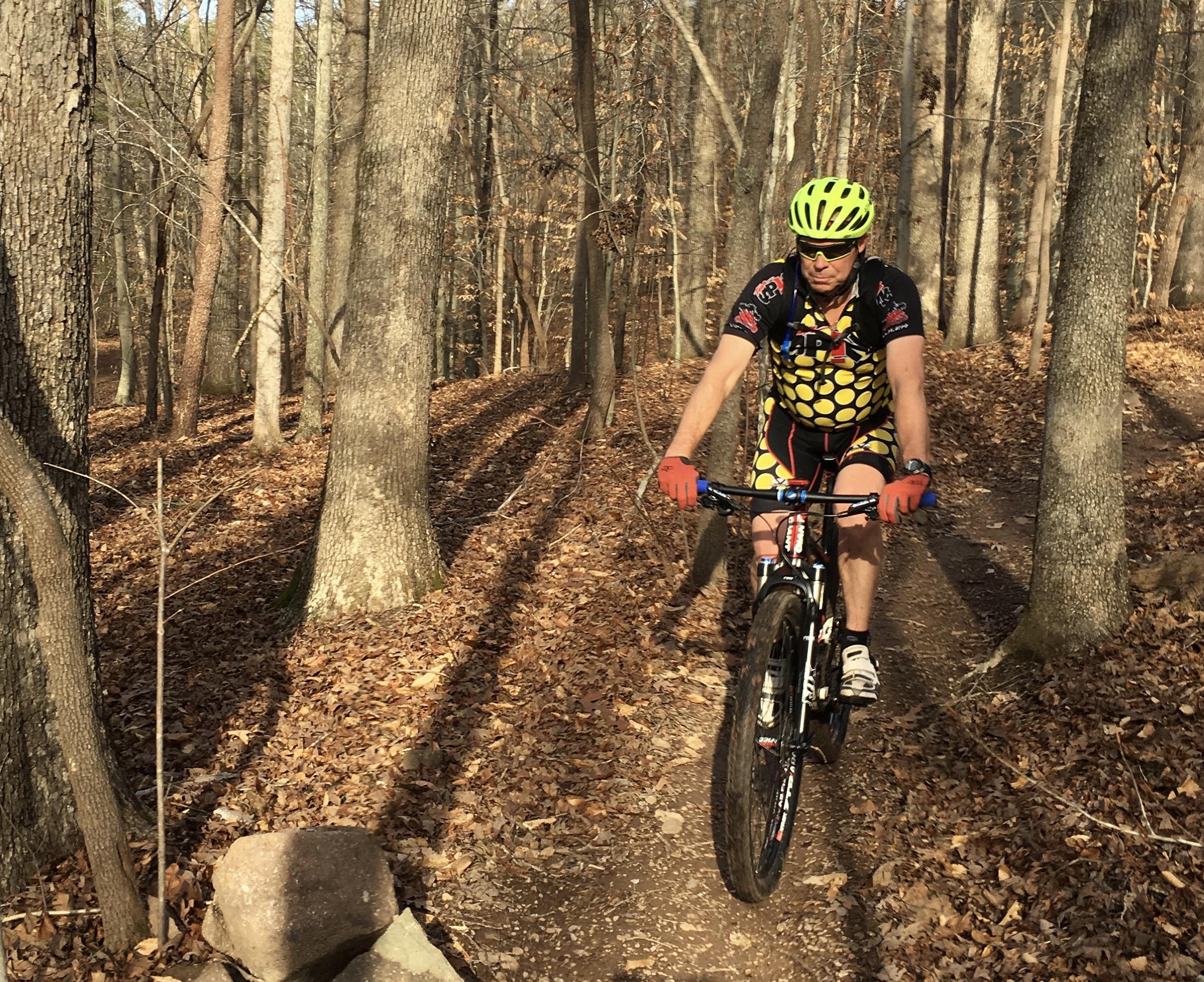 A cyclist riding a mountain bike on a leaf-covered trail surrounded by tall trees in a forest. The rider is wearing a brightly colored, patterned jersey and a yellow helmet, with a focused expression as they navigate the terrain. Sunlight filters through the trees, creating a warm atmosphere. Farris Park mountain bike trail.