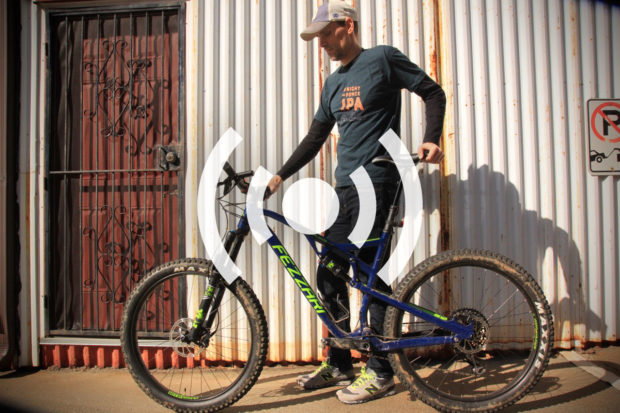 A person standing next to a mountain bike on a sunny day, with a rustic, corrugated metal wall and a brown door in the background. The individual is wearing a dark t-shirt and a cap, smiling as they hold the bike's handlebars.