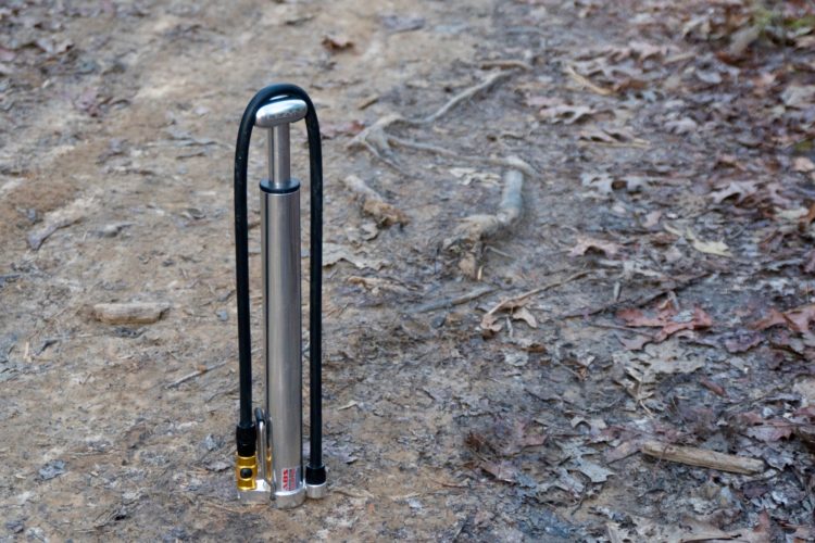 A vertical stainless steel pump with black hoses, standing on a dirt path covered in leaves and twigs. The pump has two connections at its base, indicating it's designed for a specific function, likely related to water or air.
