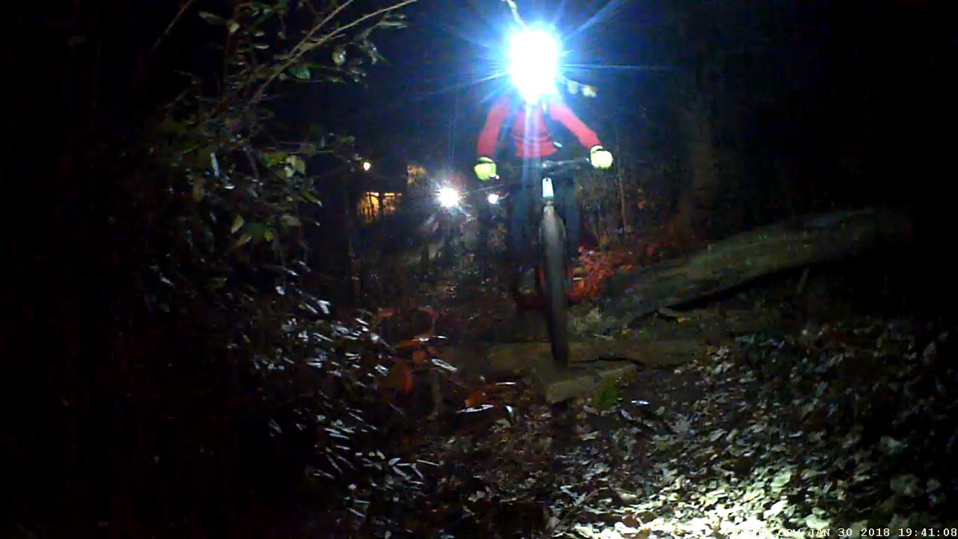 A cyclist riding on a dimly lit path at night, illuminated by a bright headlamp. The surrounding area features dark foliage and a fallen log, creating a natural trail atmosphere. Another light source can be seen in the background, indicating additional cyclists in the area. Waterworks / Mason Mill mountain bike trail.