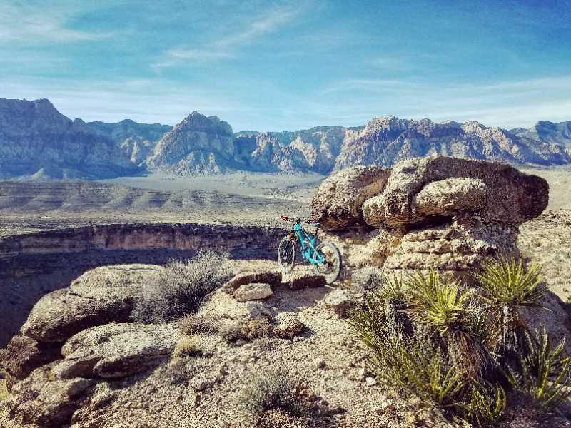 A mountain bike resting on a rocky outcrop with a vast desert landscape and mountains in the background, under a clear blue sky. Sparse vegetation and rugged terrain are visible in the foreground. Cowboy Trails mountain bike trail.