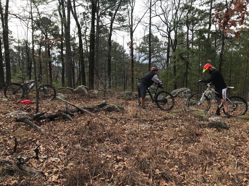 Two mountain bikers adjusting their bikes on a wooded trail. In the background, two bicycles are parked among rocks and fallen leaves, with trees and shrubs surrounding the area. The setting appears to be a natural, outdoor environment. Little Blakley mountain bike trail.