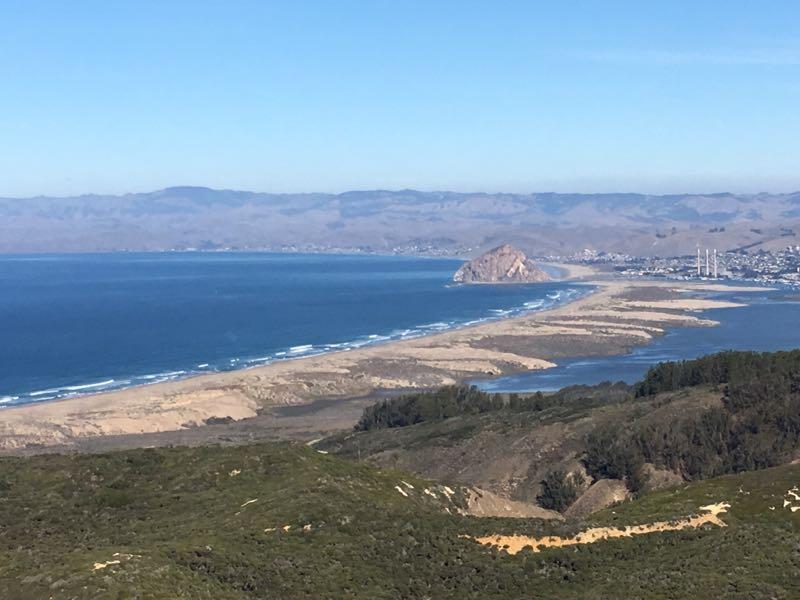 A scenic view of a coastal landscape featuring a sandy peninsula extending into the ocean, with gentle waves lapping at the shore. In the background, rolling hills and distant mountains are visible under a clear blue sky. A prominent rock formation can be seen on the peninsula, with a small town and power plant in the distance. The foreground includes lush green vegetation. Montana De Oro mountain bike trail.
