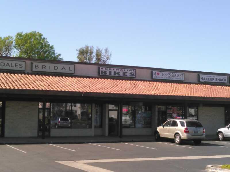 A storefront in a shopping center featuring various businesses. Visible signs include "Aldales Bridal," "Adrenaline Bikes," "Mother's Secret," and "Makeup Shack." The building has a tiled roof and a parking lot in front, with a few parked cars. Trees are visible in the background under a clear blue sky.