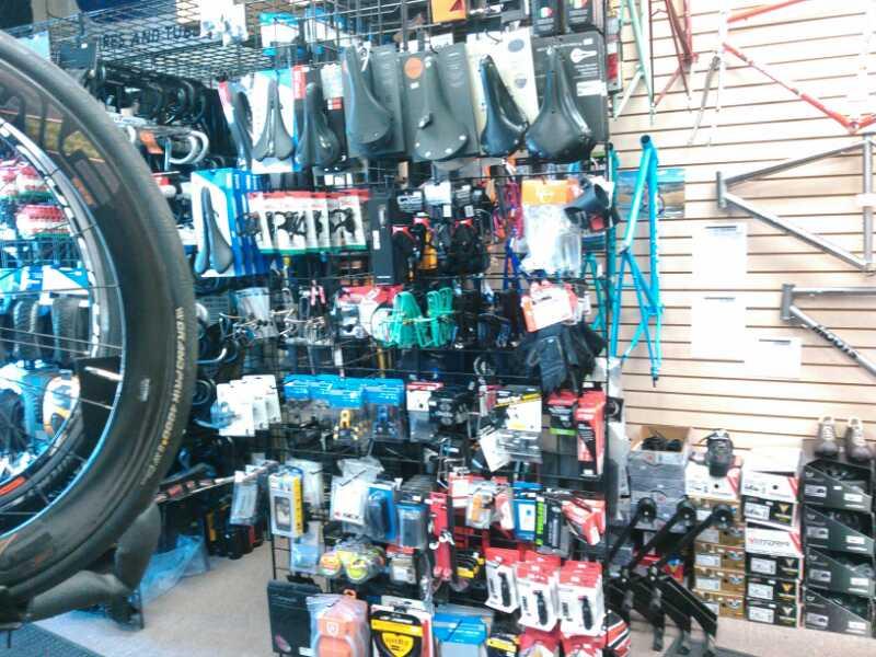 A cluttered display wall in a bike shop showcasing various cycling accessories and parts, including bike seats, tools, and safety gear, with a bicycle tire partially visible in the foreground.