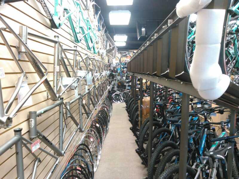 A narrow aisle in a bike shop, lined with bicycle frames displayed on the wall and various bike wheels on the floor. Several bicycles are stacked on the right side, and overhead, a rack holds additional items. The shop has bright ceiling lights illuminating the space.