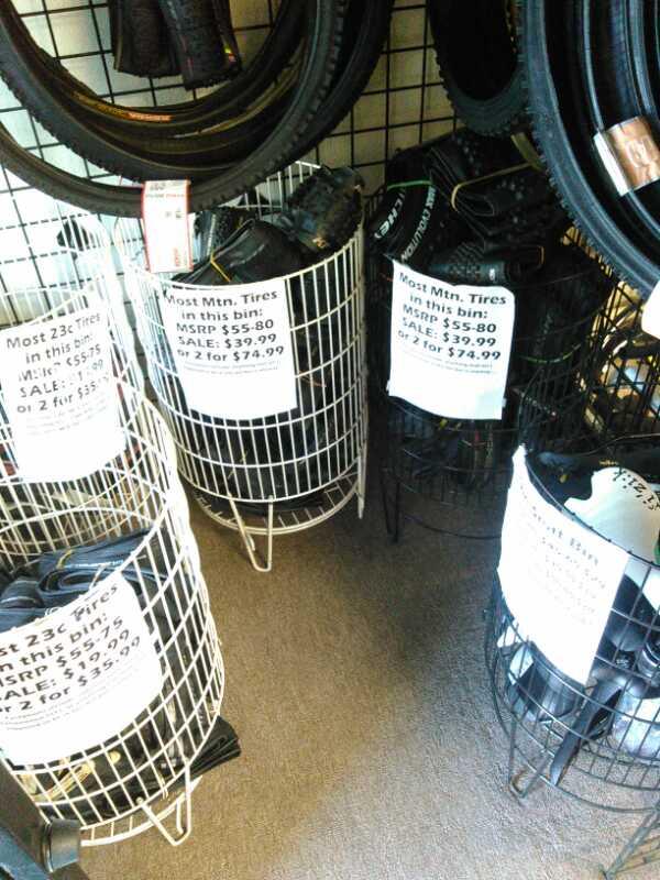 Display of bicycle tires in wire bins, each with signs listing pricing details. Bins contain mountain tires with various labels indicating MSRP and sale prices, including options for purchasing two tires at a discounted rate. The background shows additional tires hanging on a display rack.