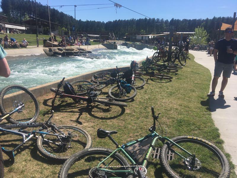A sunny outdoor scene featuring several mountain bikes parked on grassy land near a recreational water area with a flowing current. People are seen enjoying the space, with trees lining the background and amenities in sight. USNWC mountain bike trail.
