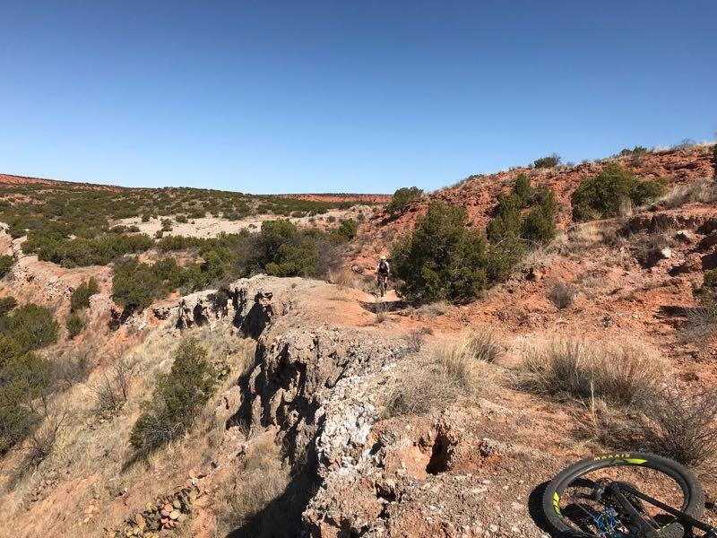 A mountain biker navigating a narrow trail along rocky terrain in a desert landscape, with red hills and sparse vegetation under a clear blue sky. A bicycle wheel is partially visible in the foreground. Caprock Canyons Trail System mountain bike trail.