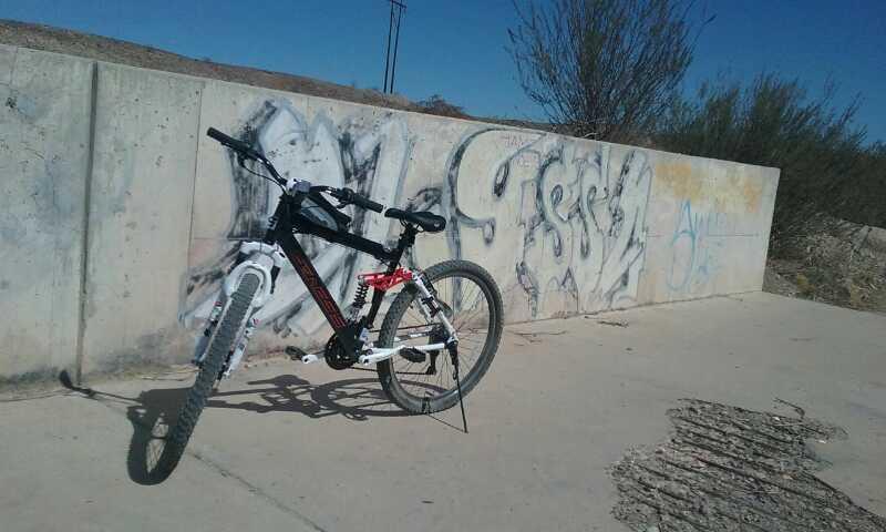 A black and white mountain bike leaning against a concrete wall covered with graffiti. The background features a clear blue sky and sparse vegetation. The ground shows evidence of wear, with visible cracks and markings. Sugarloaf Peak mountain bike trail.