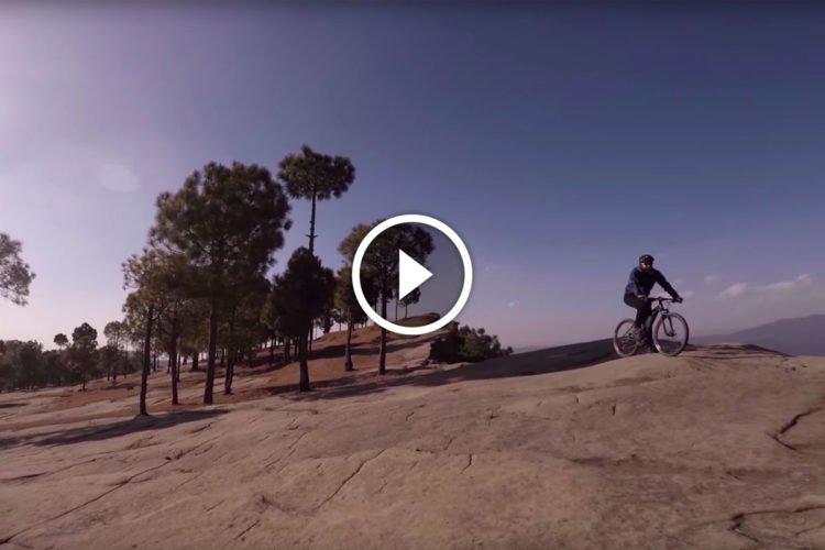 A cyclist riding on a rocky landscape surrounded by trees and a clear blue sky. The scene captures a sense of adventure and outdoor activity.