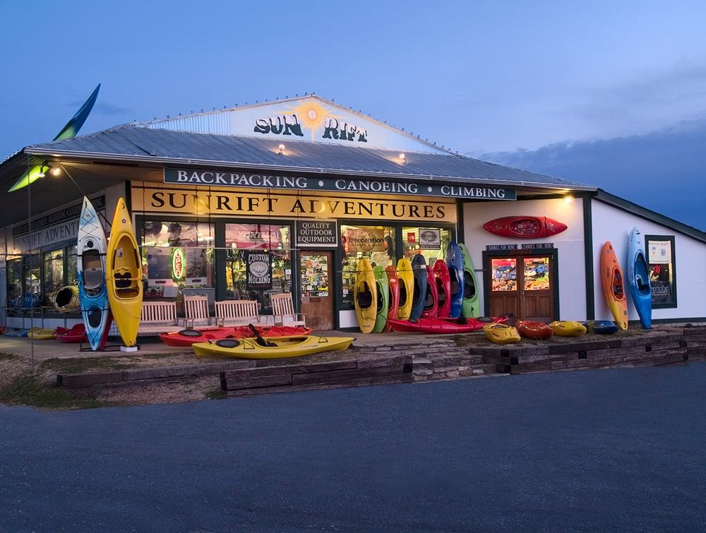 A storefront for Sun Rift Adventures, featuring a variety of colorful kayaks displayed outside. The building has a large sign and showcases outdoor equipment for backpacking, canoeing, and climbing. The scene is set during dusk, with warm lighting illuminating the shop.