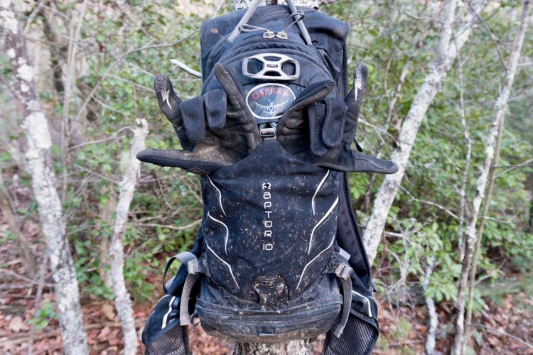 A black Osprey Raptor 10 backpack hanging from a tree branch, with a pair of muddy gloves positioned in a playful manner on the front of the backpack. The background features greenery and tree trunks, suggesting an outdoor, forested environment.