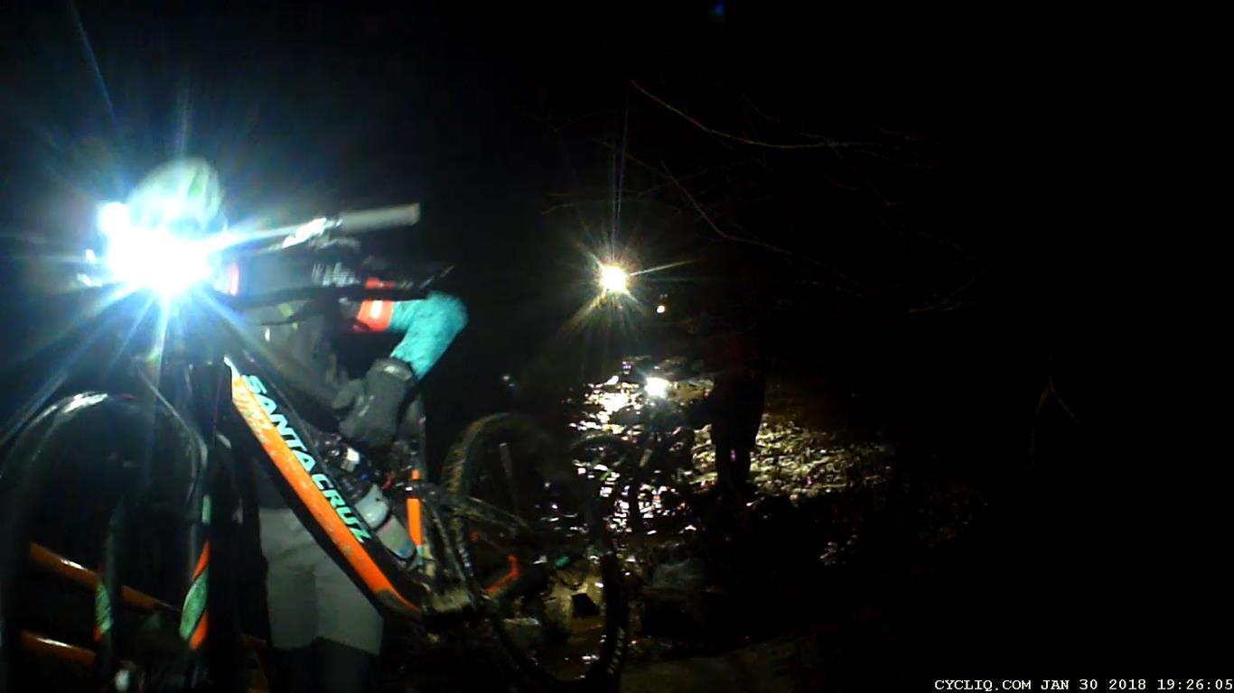 A cyclist maneuvering a bike through a dark, muddy trail, illuminated by bright bike lights. Another cyclist is visible in the background, also navigating the path. The scene depicts a nighttime biking adventure in a rugged environment. Waterworks / Mason Mill mountain bike trail.