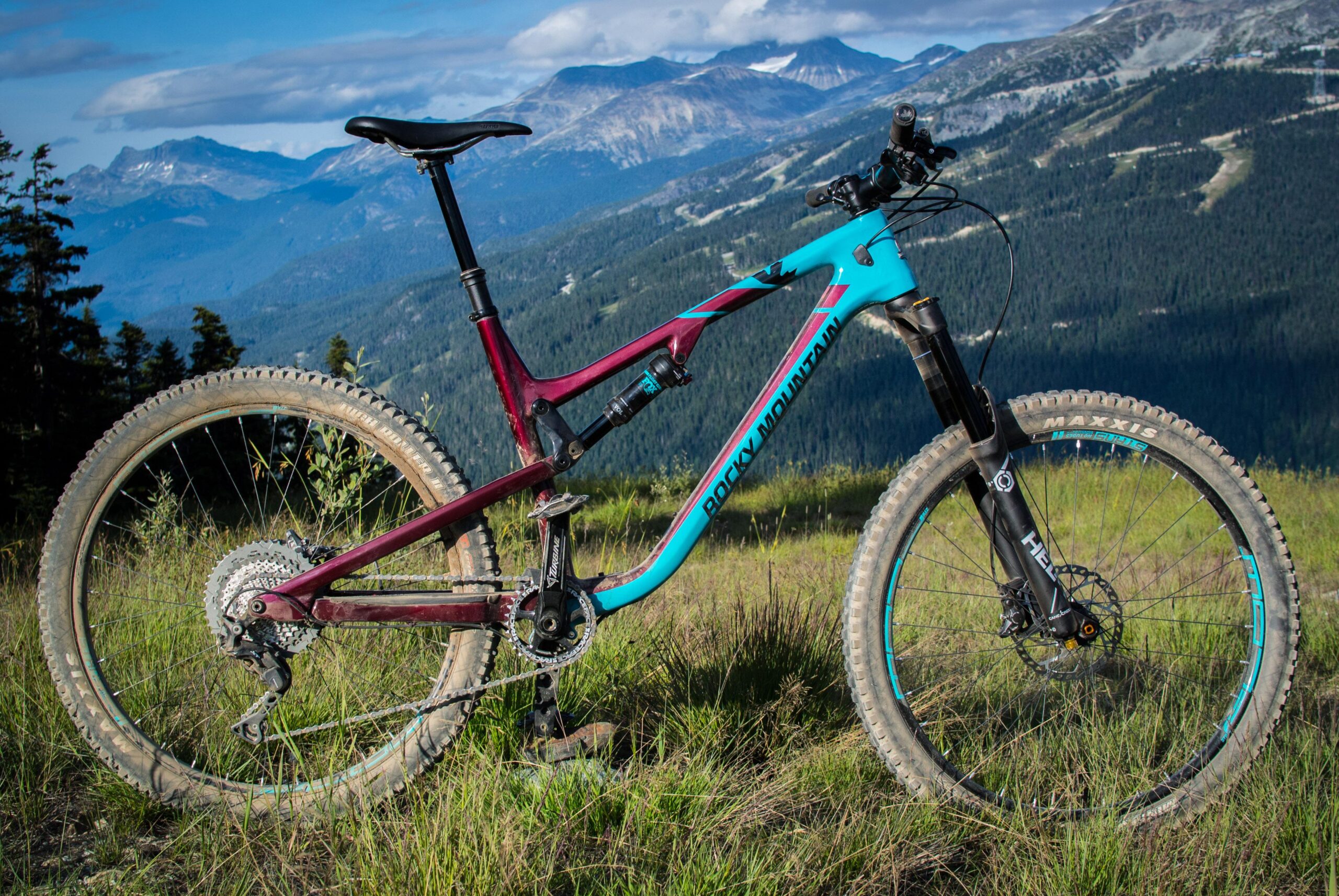 Rocky Mountain Altitude 750: A mountain bike resting on grass with a scenic backdrop of mountains and trees. The bike features a maroon and turquoise frame, rugged tires, and a mountain bike setup suitable for trails. The surrounding landscape includes lush greenery and distant peaks under a cloudy sky.