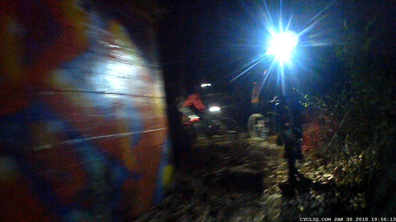 A dimly lit scene at night featuring a colorful, graffiti-covered wall on the left side. In the background, there are faint silhouettes of individuals near vehicles illuminated by bright lights. The environment appears to be an outdoor area with foliage and uneven ground. Waterworks / Mason Mill mountain bike trail.