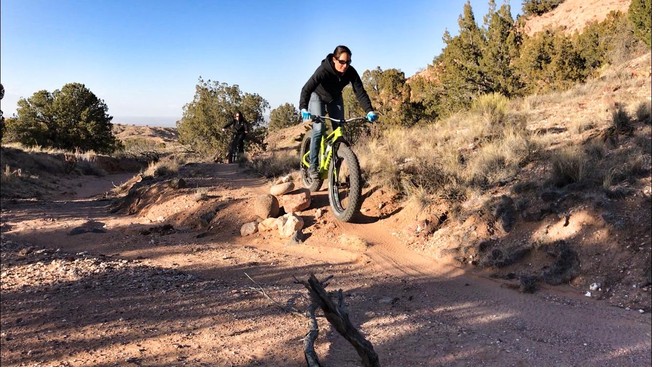 A cyclist riding a fat bike over rough terrain on a sunny day. The bike is bright green, and the rider is wearing a black hoodie and gloves. In the background, another person can be seen biking on a dirt path surrounded by sparse vegetation and rocky landscapes. Mariposa Fat Bike Trails mountain bike trail.