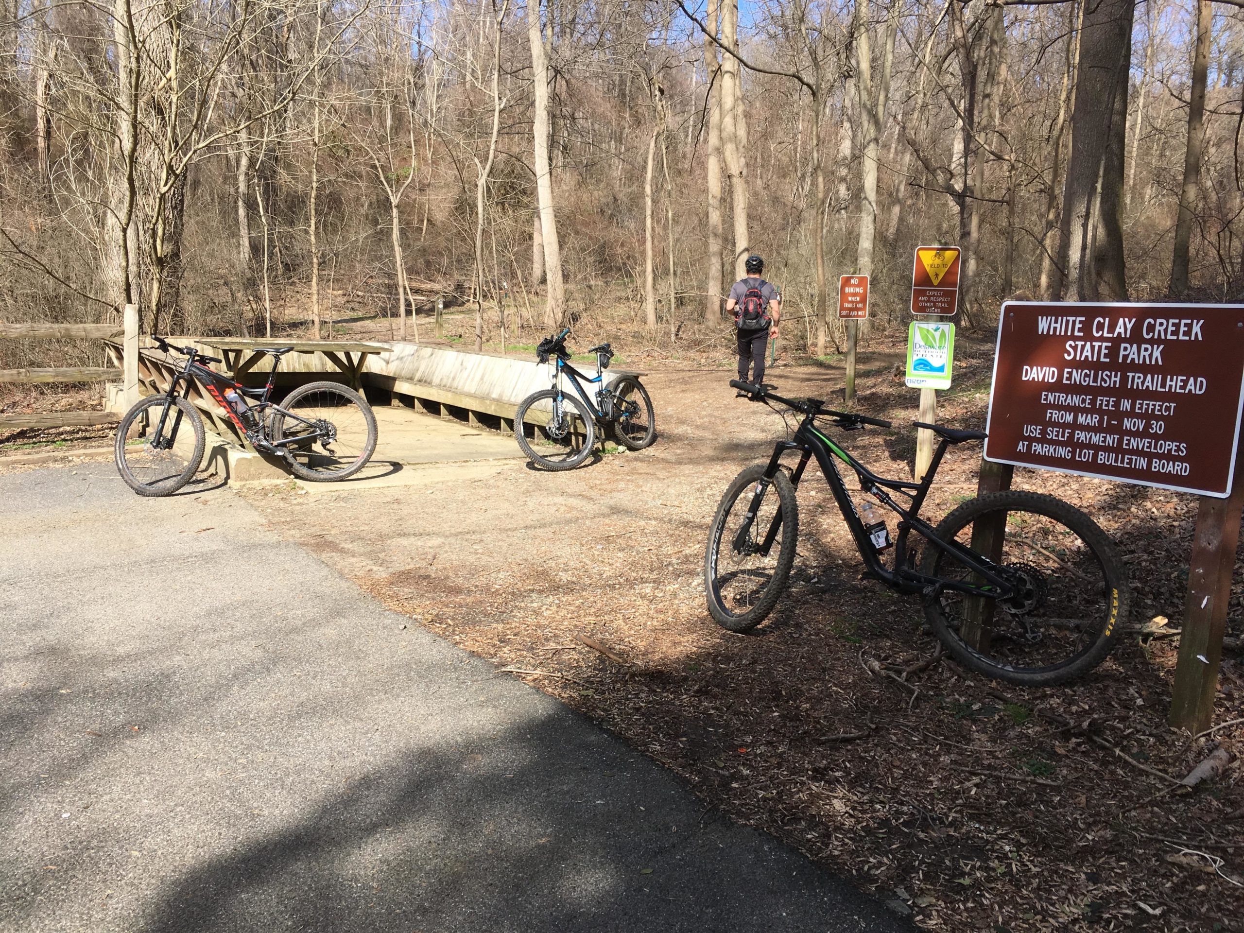 A scenic view of the David English Trailhead at White Clay Creek State Park, featuring three mountain bikes parked on the ground near a wooden bridge. A person is seen walking on the trail, and several informational signs are visible, indicating park rules and entrance fees. The surrounding area is wooded with bare trees, suggesting early spring or late winter. White Clay Creek mountain bike trail.