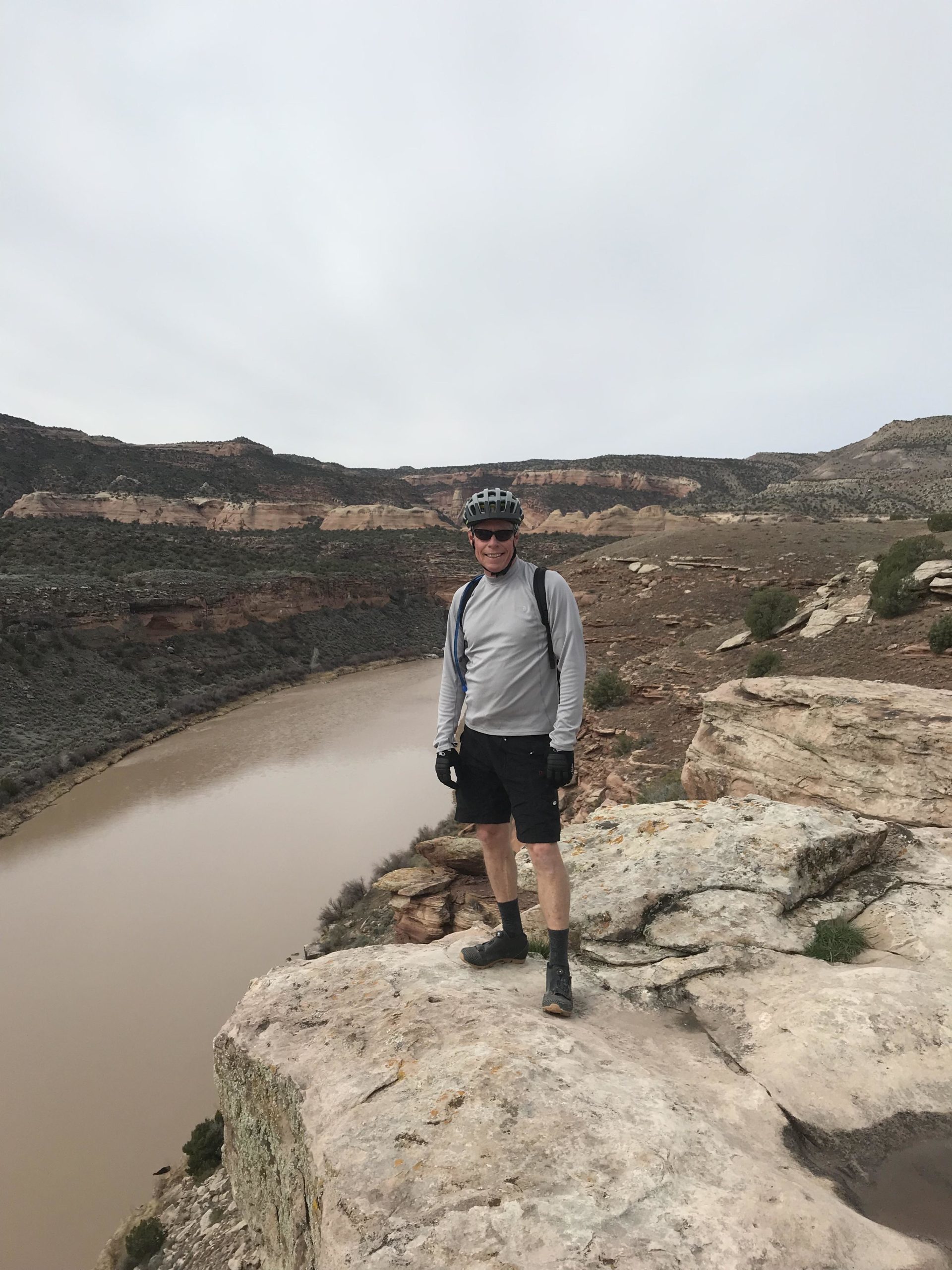 A person wearing a helmet and sunglasses stands on a rocky ledge overlooking a river. The landscape features rugged hills and cliffs in the background, with a cloudy sky above. The individual is dressed in outdoor gear, including a long-sleeve shirt and shorts, and appears to be enjoying the scenic view. Mary's Loop / Horsethief Bench mountain bike trail.