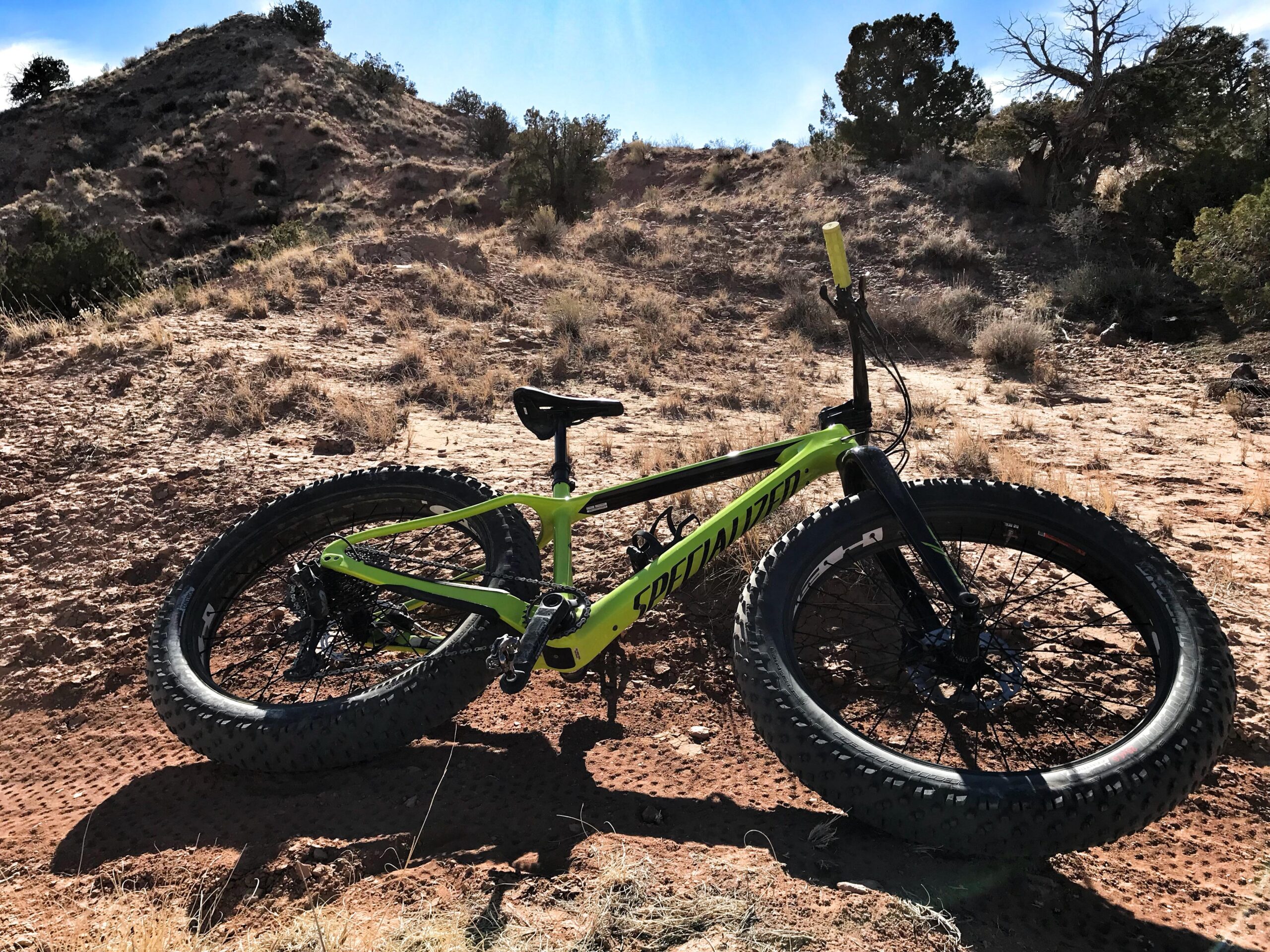 Specialized Fatboy Carbon Comp: A bright green fat tire bike resting on rocky terrain, surrounded by sparse vegetation and hills under a clear blue sky.