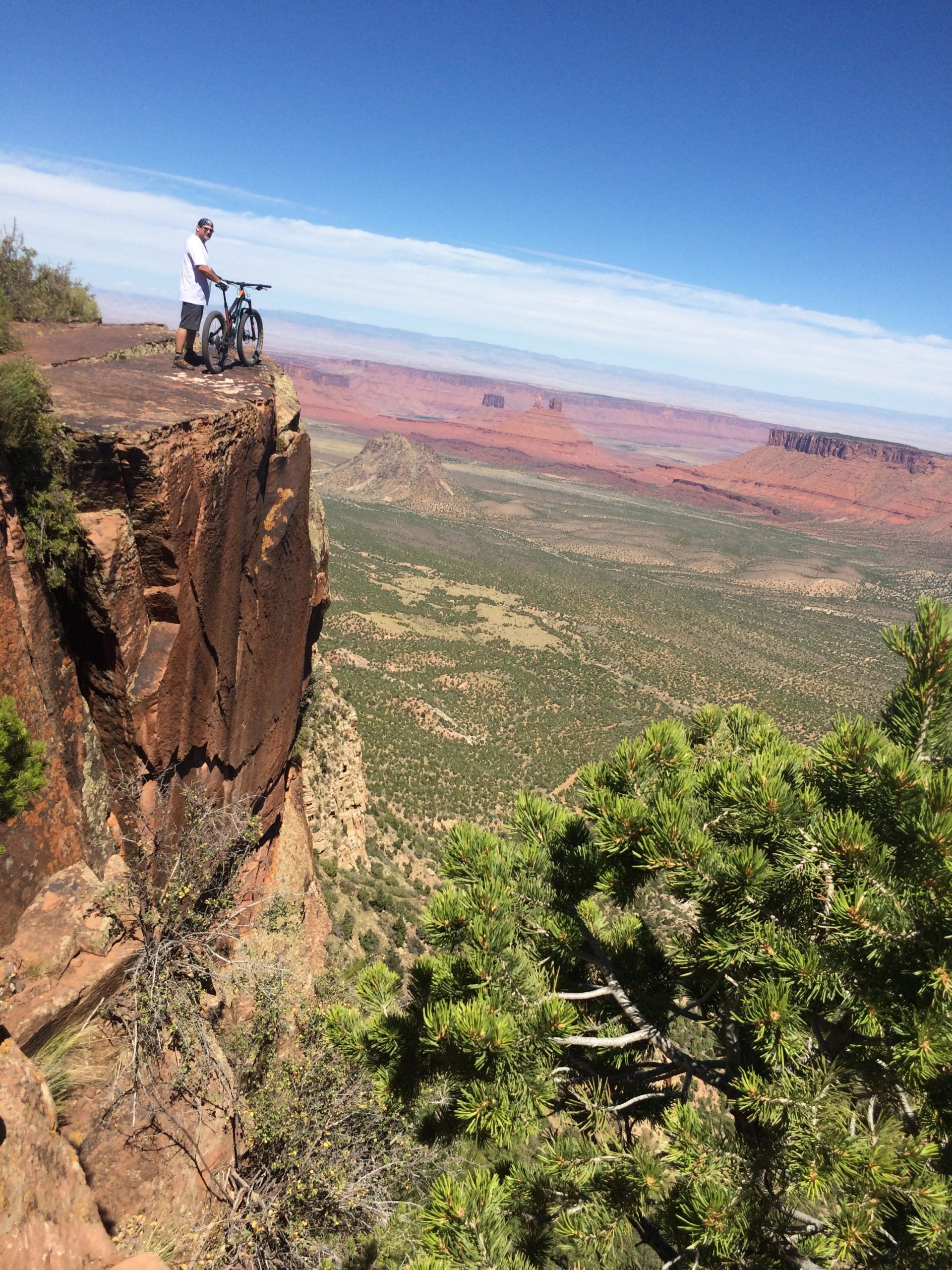 A person standing on a rocky cliff edge with a mountain bike, overlooking a vast landscape of red rock formations and green vegetation under a clear blue sky. The Whole Enchilada mountain bike trail.