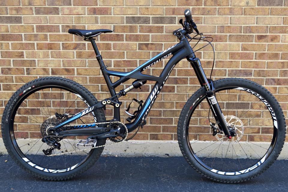 Specialized Enduro Elite: A black and blue mountain bike with a sleek design, featuring thick tires and a modern frame, parked against a brick wall.