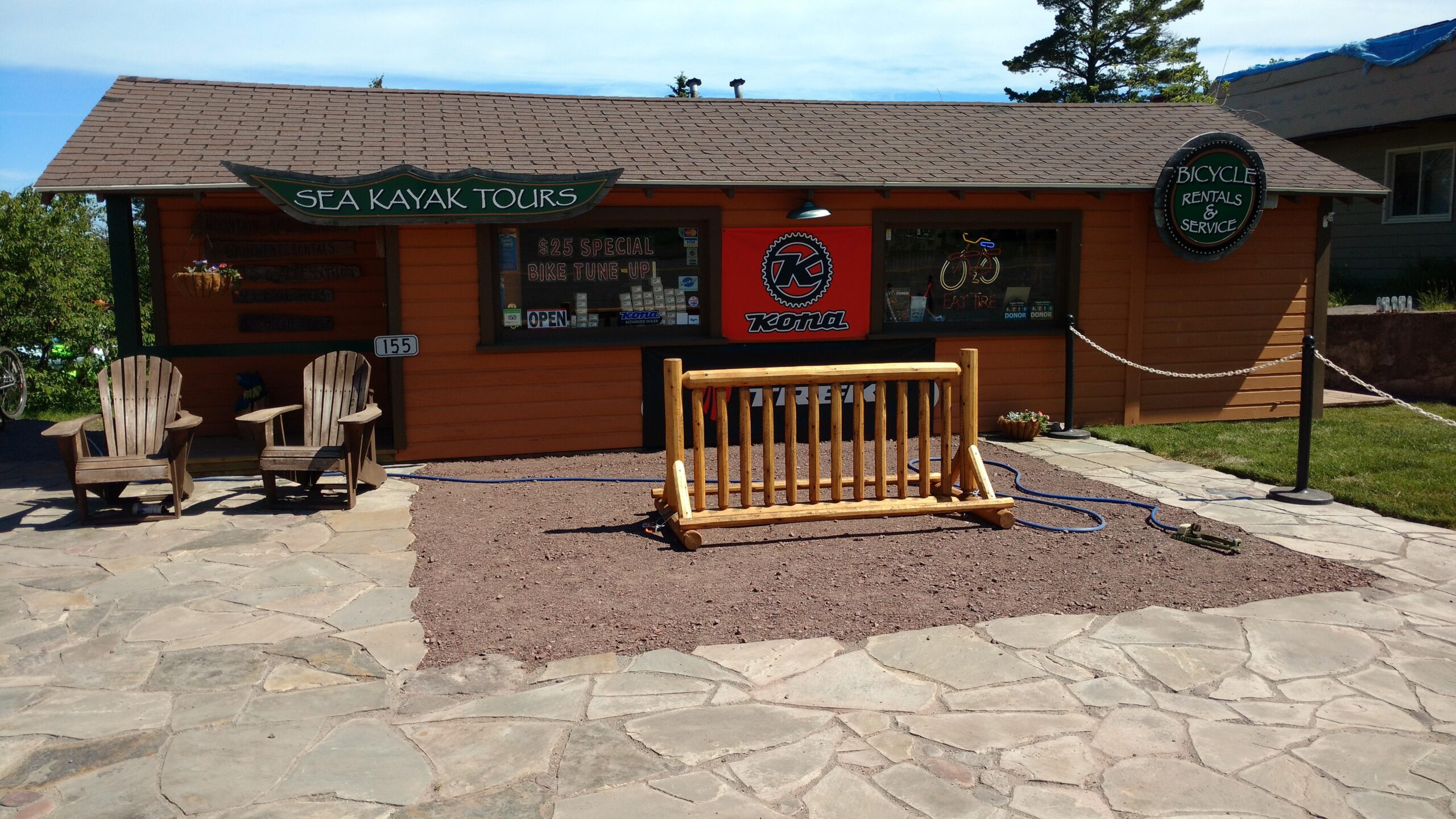 A small, charming outdoor shop offering sea kayak tours and bicycle rentals, set against a clear blue sky. The building features a wooden sign indicating "Sea Kayak Tours" and another for "Bicycle Rentals & Service." In front, there are two wooden chairs and a bike rack, surrounded by a stone pathway and landscaped grass.