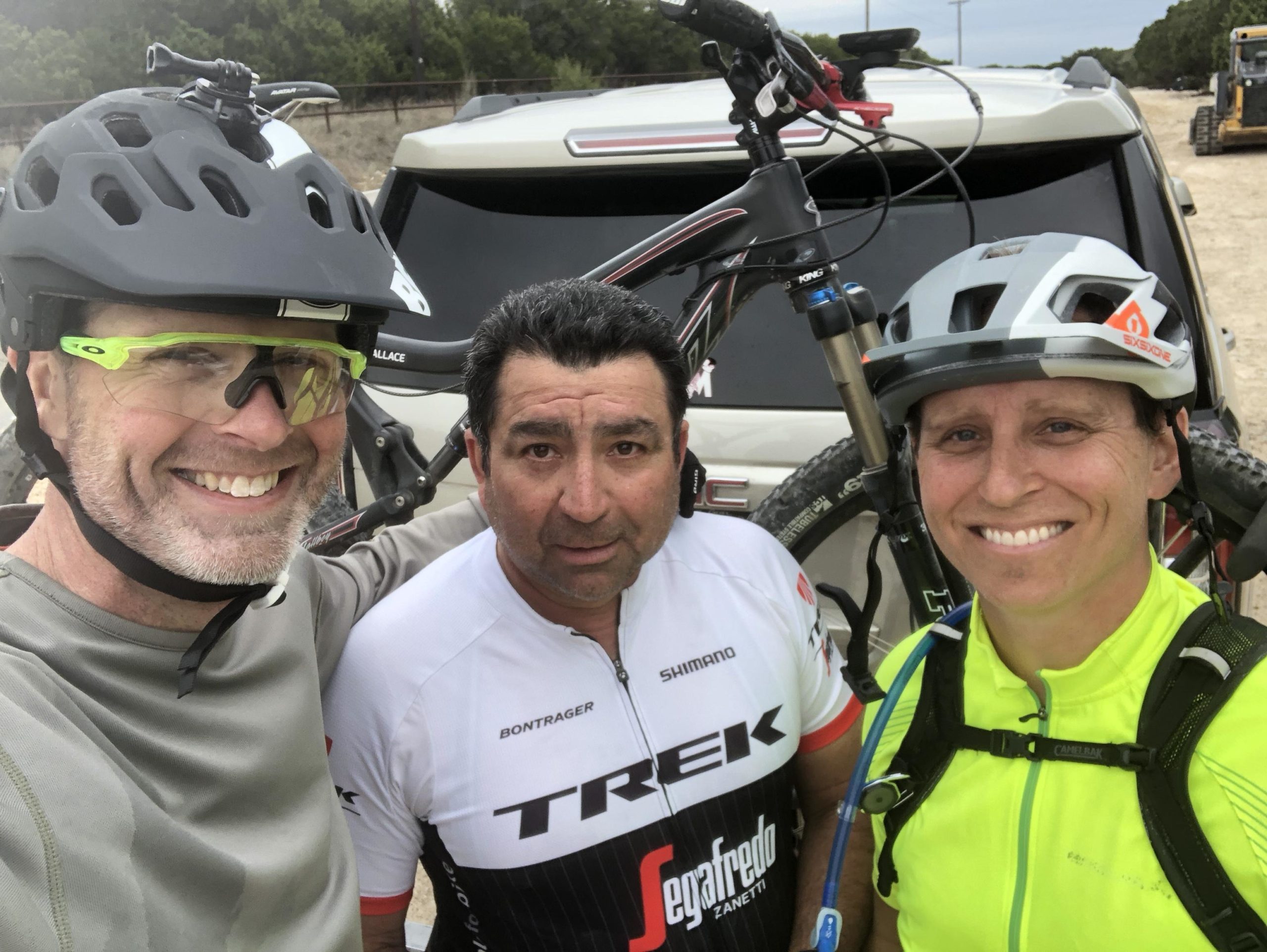Three cyclists pose for a selfie outdoors, smiling at the camera. The man on the left is wearing a gray shirt and a helmet with a camera attached, while the middle figure is in a white and black biking jersey. The person on the right is dressed in a bright yellow top with a hydration pack. In the background, a vehicle with mountain bikes is visible, along with trees and a gravel pathway. Dana Peak mountain bike trail.