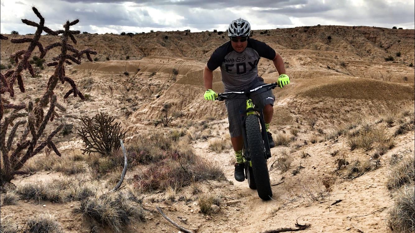 A person riding a fat tire bike on a sandy, rugged terrain surrounded by desert vegetation, including cacti and sparse grass. The biker is wearing a helmet and bright green gloves, navigating uphill against a backdrop of cloud-covered skies and rolling hills. Mariposa Fat Bike Trails mountain bike trail.