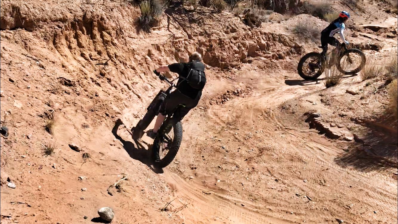 Two cyclists navigate a rugged dirt trail in a desert landscape. The first rider, wearing a backpack, is seen leaning into a turn on a fat tire bike, while the second cyclist appears in the background, approaching the same curve. The trail is surrounded by sandy soil and sparse vegetation. Mariposa Fat Bike Trails mountain bike trail.