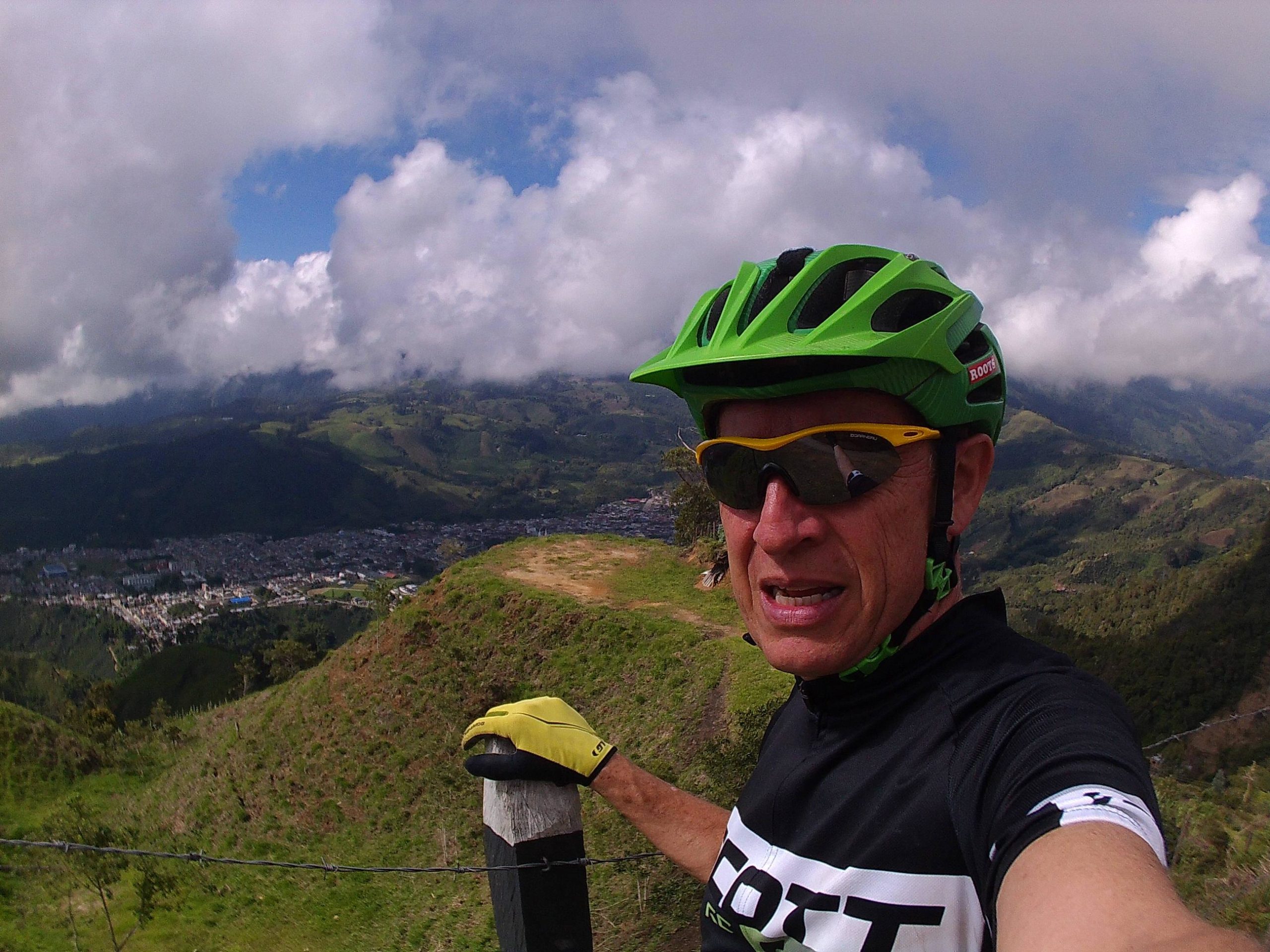 A person wearing a bright green helmet and sunglasses takes a selfie while standing on a mountain overlooking a valley. The landscape features rolling hills and a small town visible below, with a dramatic sky filled with clouds. The individual wears a black cycling jersey and holds onto a post, enjoying the scenic view. Los Nevados National Park mountain bike trail.