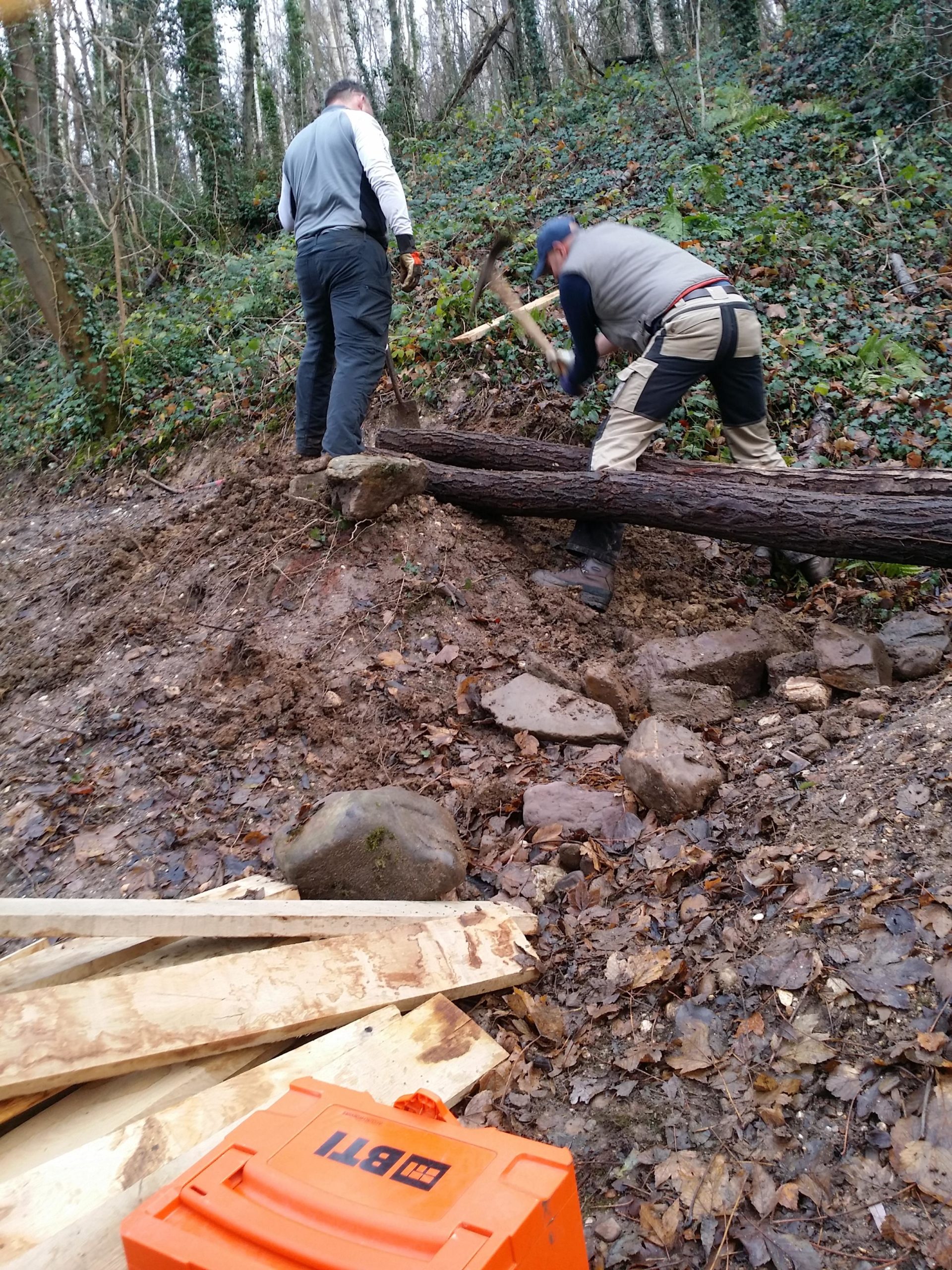 Two individuals working in a wooded area, using tools to shape the ground and move logs. A variety of rocks and wooden planks are visible in the foreground, along with an orange toolbox. The background features trees and greenery. MTB Sint Pietersbergroute mountain bike trail.