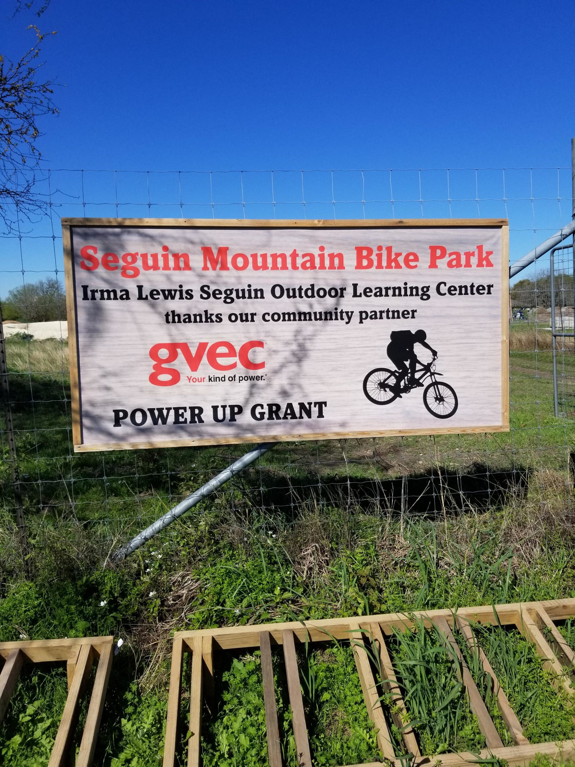 Sign at Seguin Mountain Bike Park displaying the name of the park, a note of thanks to the community partner, GVEC, and information about the Irma Lewis Seguin Outdoor Learning Center and the "Power Up Grant." The background features a clear blue sky and green grass, with wooden structures partially visible at the bottom of the image. Seguin Outdoor Learning Center mountain bike trail.