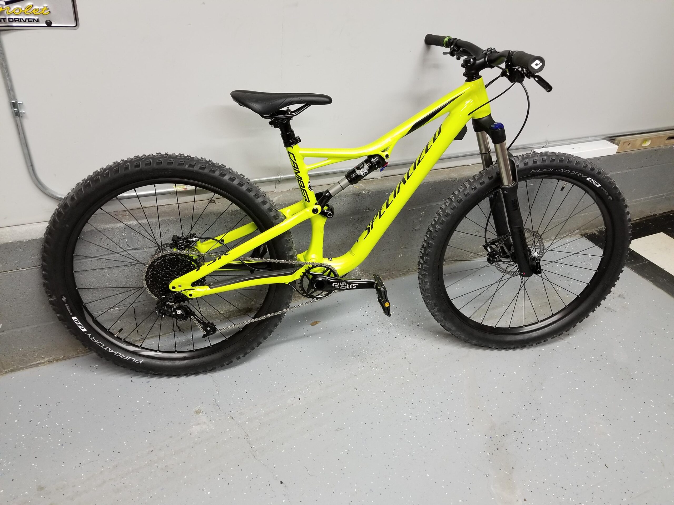 Specialized Camber: A bright yellow mountain bike is leaning against a wall in a garage. The bike features thick tires suitable for off-road terrain, a modern frame design, and hydraulic disc brakes. The floor is a grey epoxy finish, and there are visible electrical wires and a small section of wall near the bike.