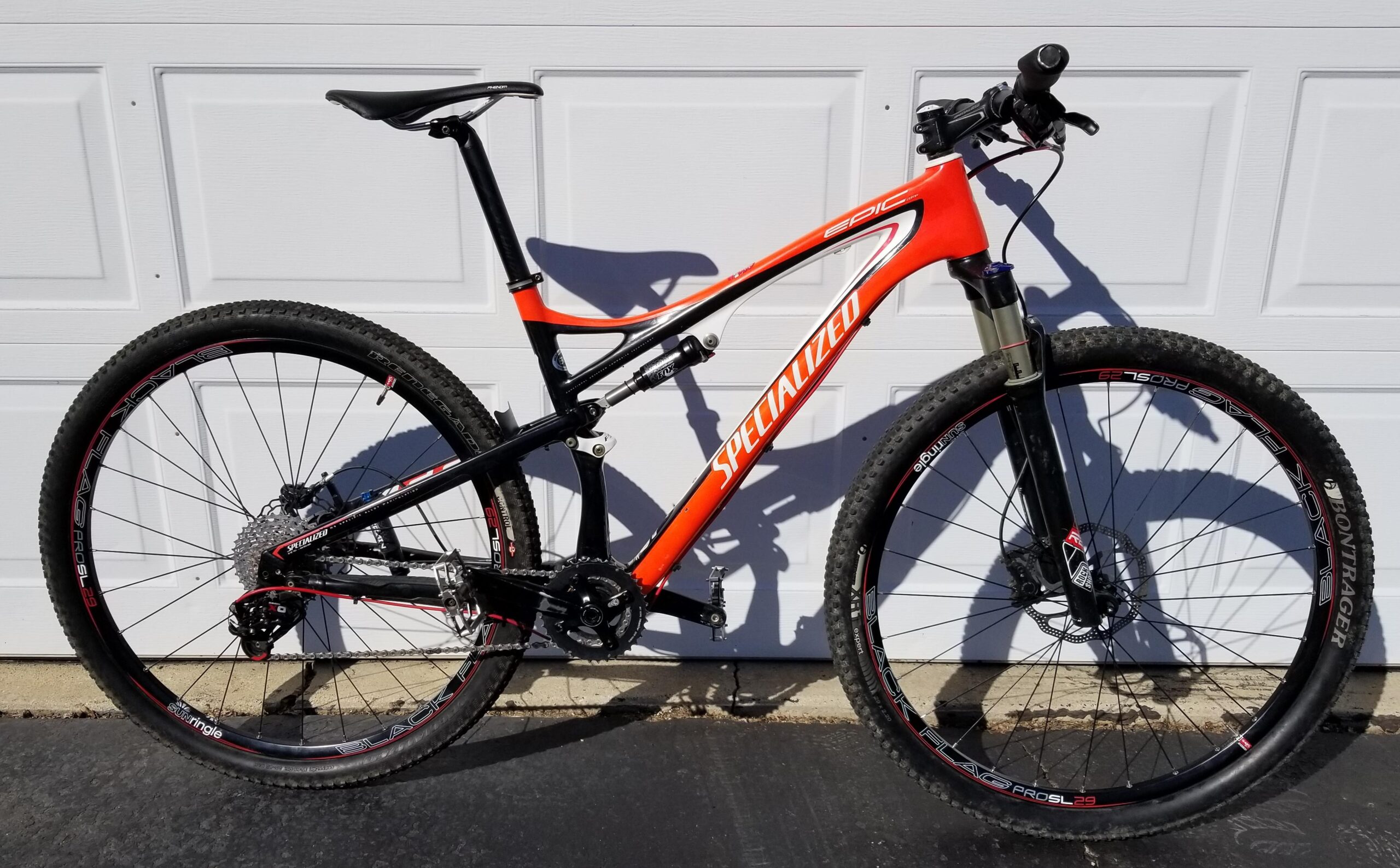 Specialized Epic Carbon Comp: An orange and black Specialized Epic mountain bike is parked next to a white garage door. The bike features thick tires suited for off-road terrain, a suspension fork, and a sleek frame design, emphasizing its performance and agility.