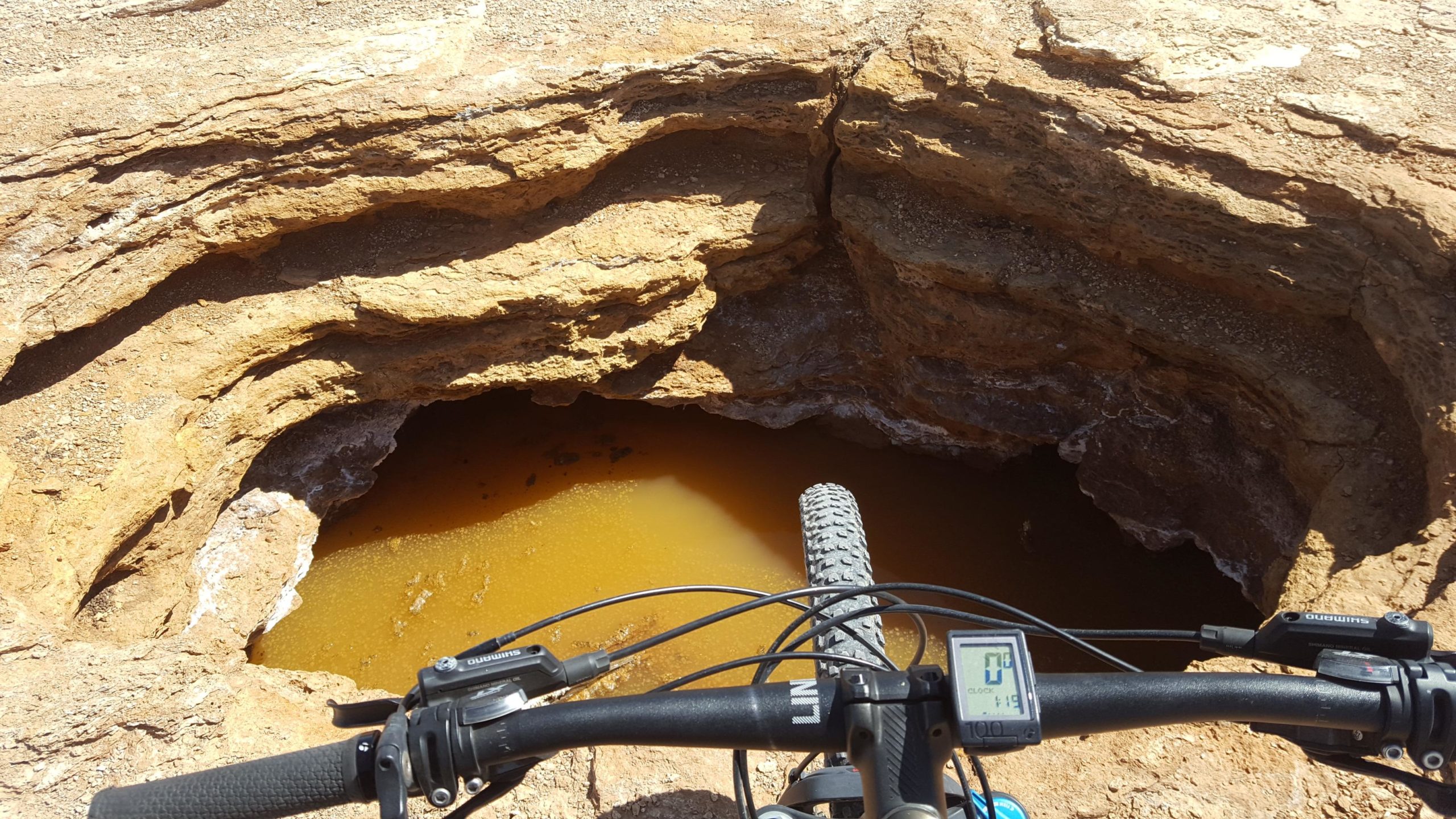 A view from above a large, deep pit in the ground, showing some stagnant brown water at the bottom. The image includes a portion of a mountain bike handlebar and a digital display, indicating the bike is stationary. The surrounding terrain is rocky and dry, with visible layers of soil and rock formations. White Ridge Bike Trails mountain bike trail.