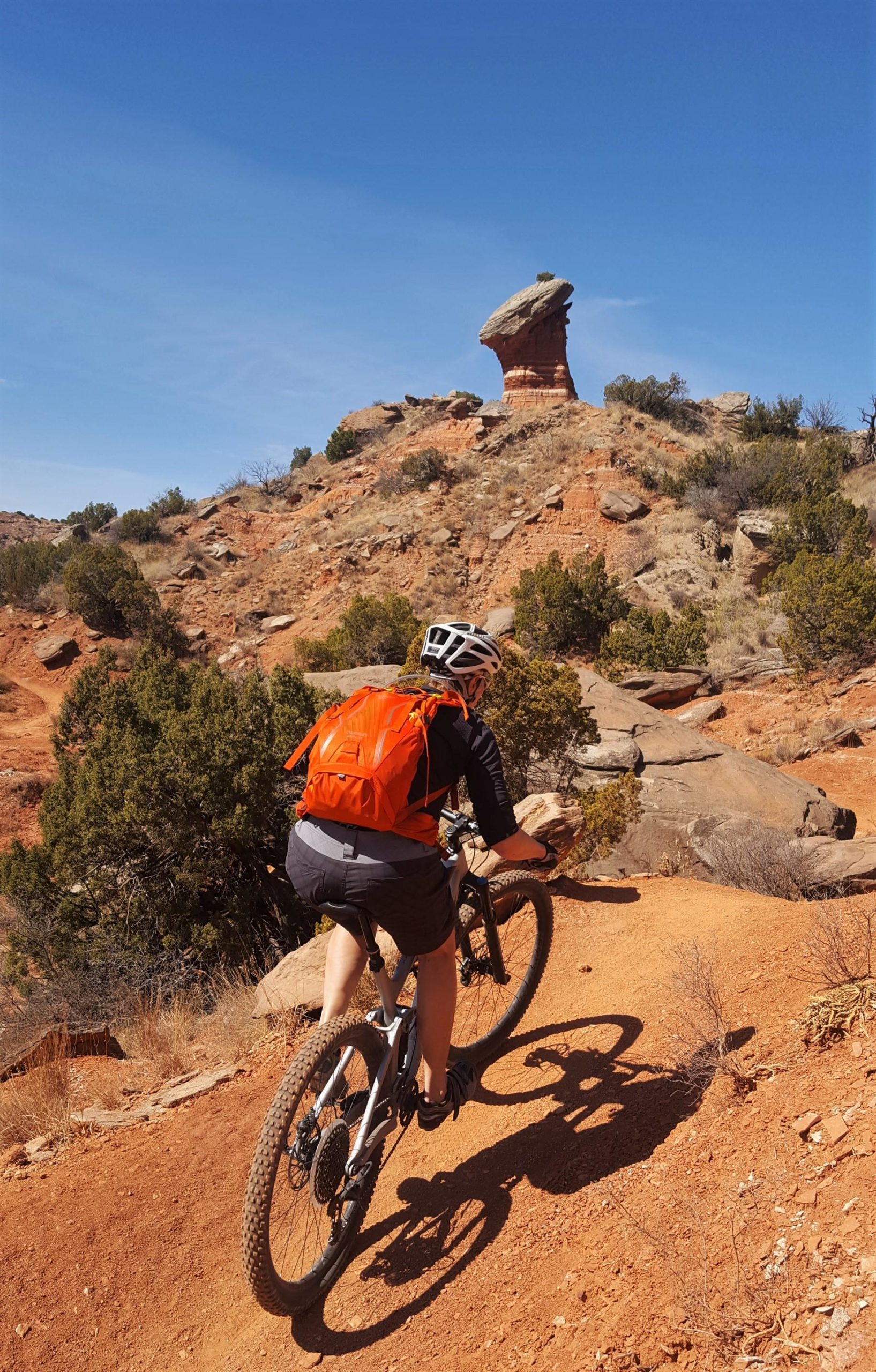 A mountain biker rides along a dirt path with rocky terrain, featuring a distinctive rock formation in the background under a clear blue sky. The biker is wearing a helmet and an orange backpack, navigating the trail surrounded by dry vegetation and shrubs. Palo Duro Canyon mountain bike trail.