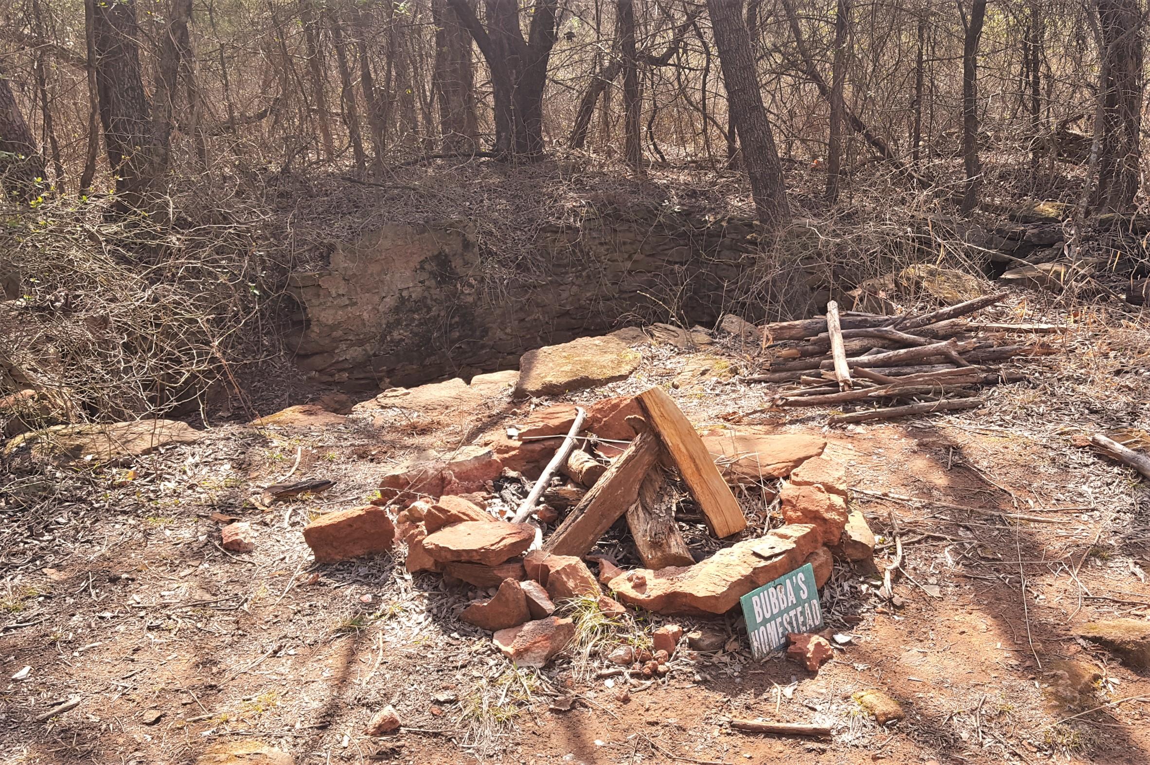 A rocky clearing surrounded by trees and underbrush, featuring a small fire pit made of stones. Next to the pit, there is a wooden sign that reads "Bubba's Homestead." Sticks and branches are stacked nearby, contributing to the rustic atmosphere of the area. Lake Stanley Draper mountain bike trail.