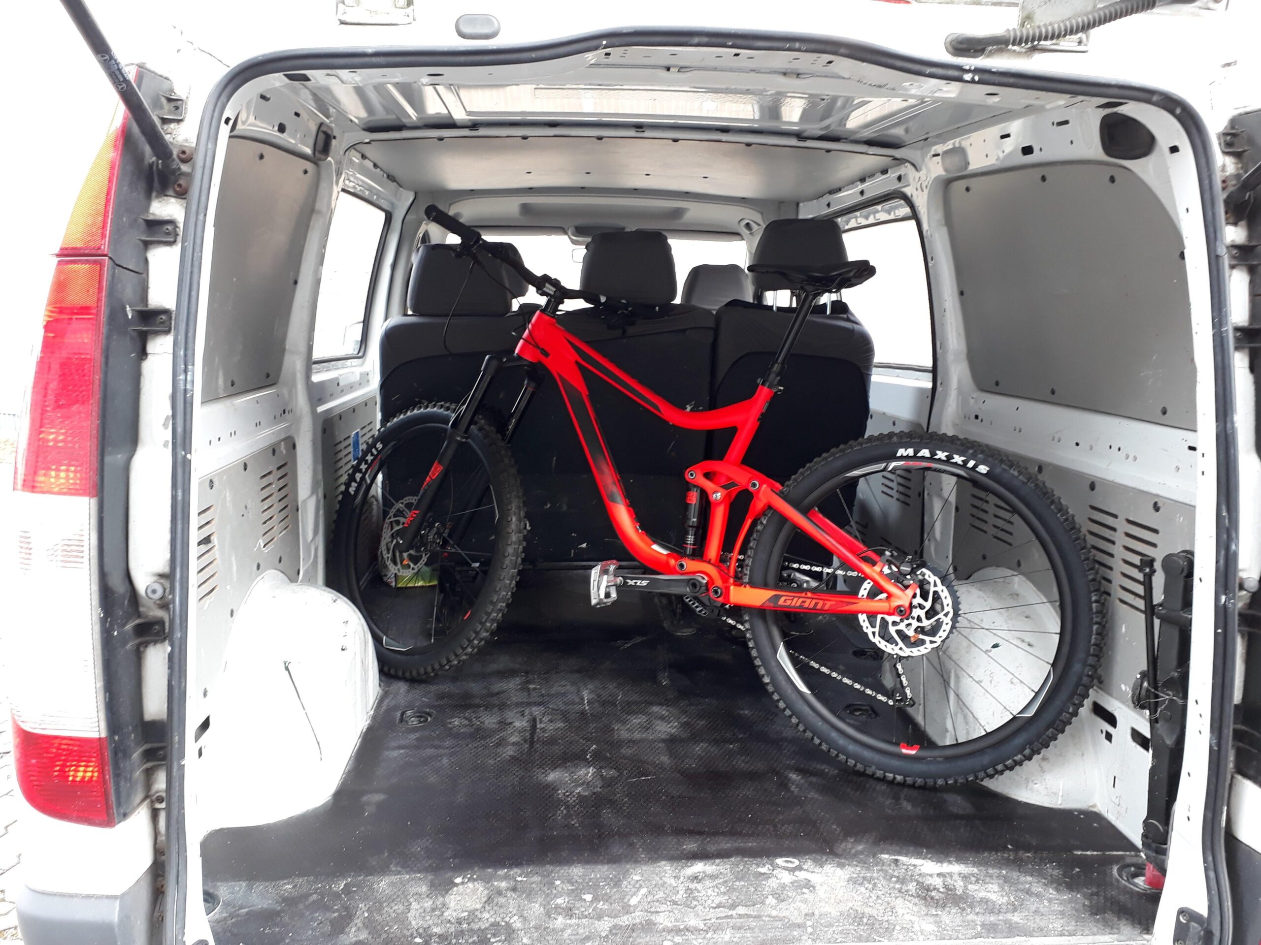 Giant Reign: A red mountain bike is secured inside the cargo area of a van, with the back doors open. The interior is spacious and features black seating in the front, while the bike is positioned upright against the side wall. The van's interior is predominantly white and gray, with a clear view of the van's cargo space.