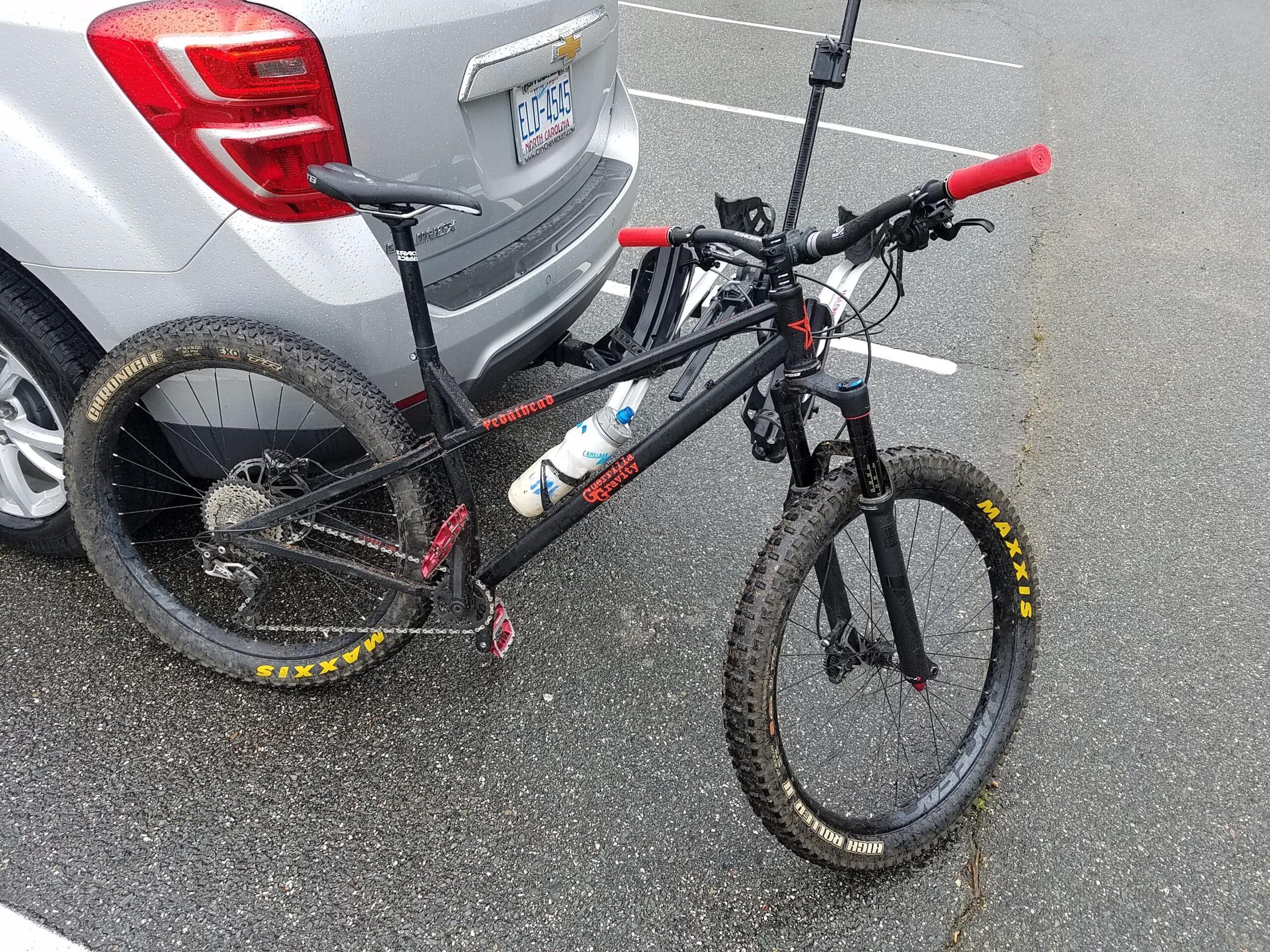 Guerrilla Gravity Pedalhead: A black mountain bike with red handlebars and muddy tires, parked next to a silver SUV in a wet parking lot. The bike is secured to a bike rack attached to the vehicle, and two water bottles are mounted on the frame.