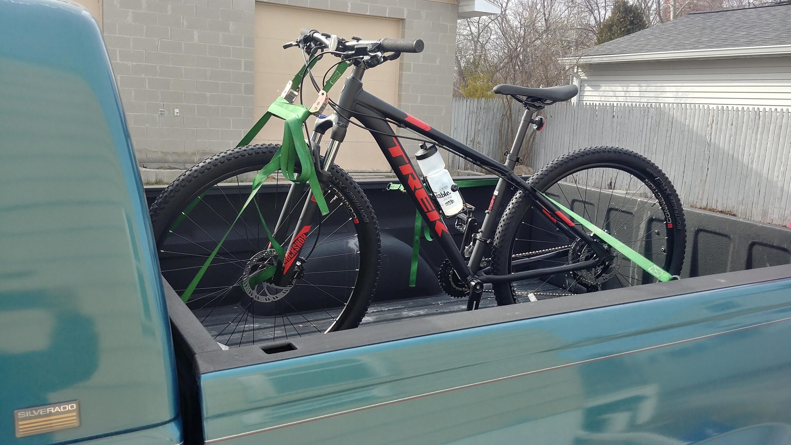 Trek Marlin 7: A black Trek mountain bike secured with green straps in the bed of a pickup truck, with a water bottle attached to the bike frame. The background features a gray wall and wooden fence, indicating an outdoor setting.