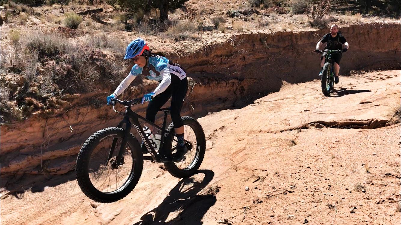 A young cyclist in a blue helmet and jersey rides over rocky terrain, while an adult on a fat bike follows closely behind. The landscape features dry dirt and sparse vegetation under a clear blue sky. Mariposa Fat Bike Trails mountain bike trail.